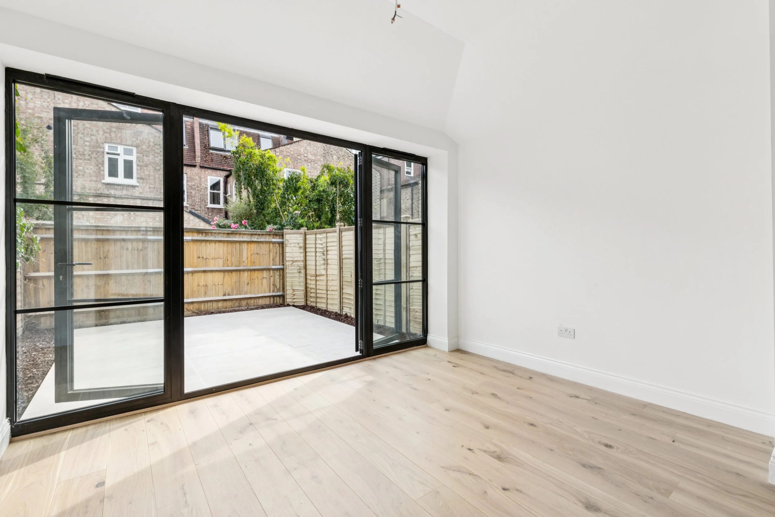 Empty room with white walls, light wood flooring, and large glass sliding doors leading to a small outdoor patio with a wooden fence and view of neighboring brick buildings and green trees.