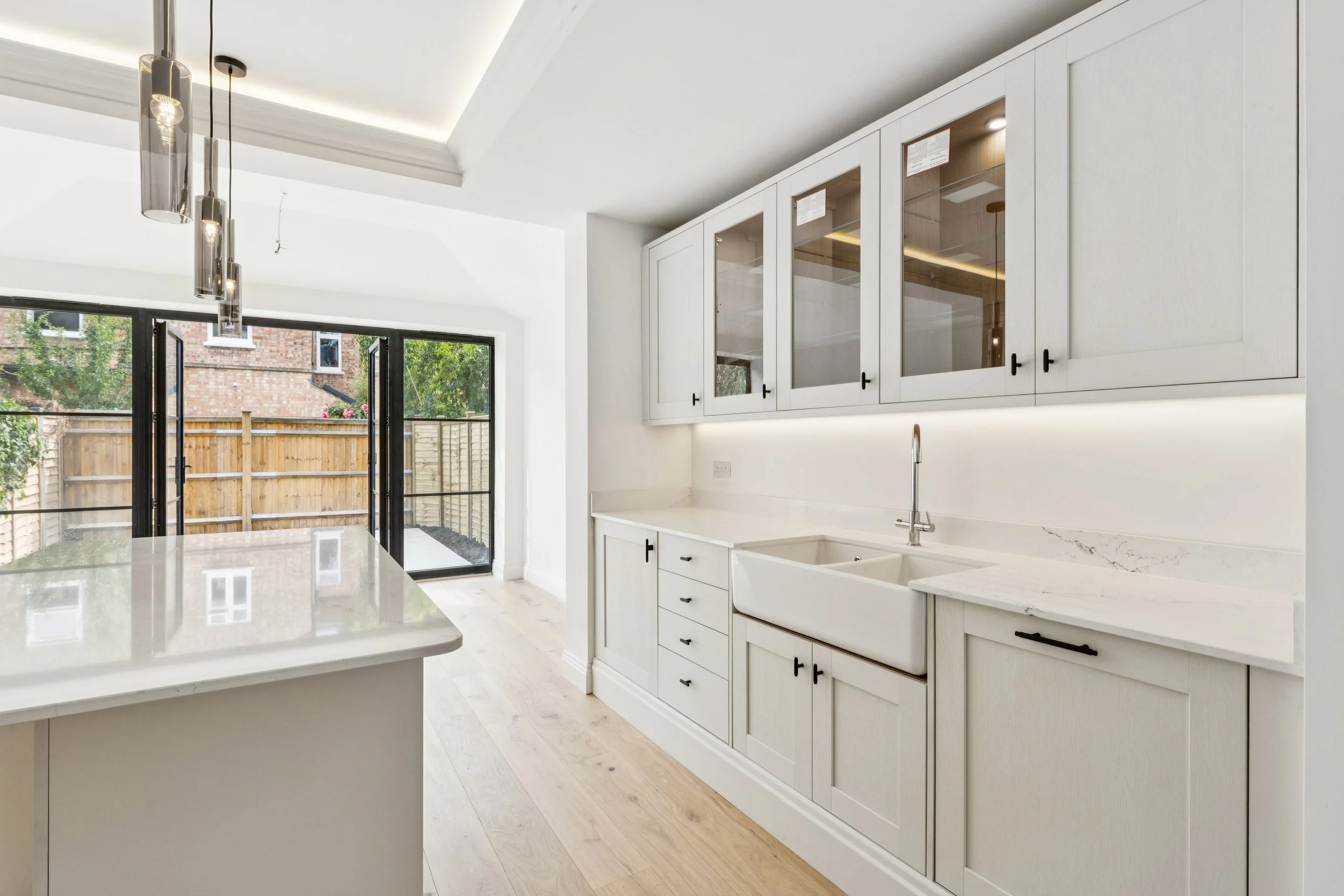 Modern white kitchen with glass-front cabinets, a white island, a double sink, and large glass doors leading to a fenced backyard with trees.