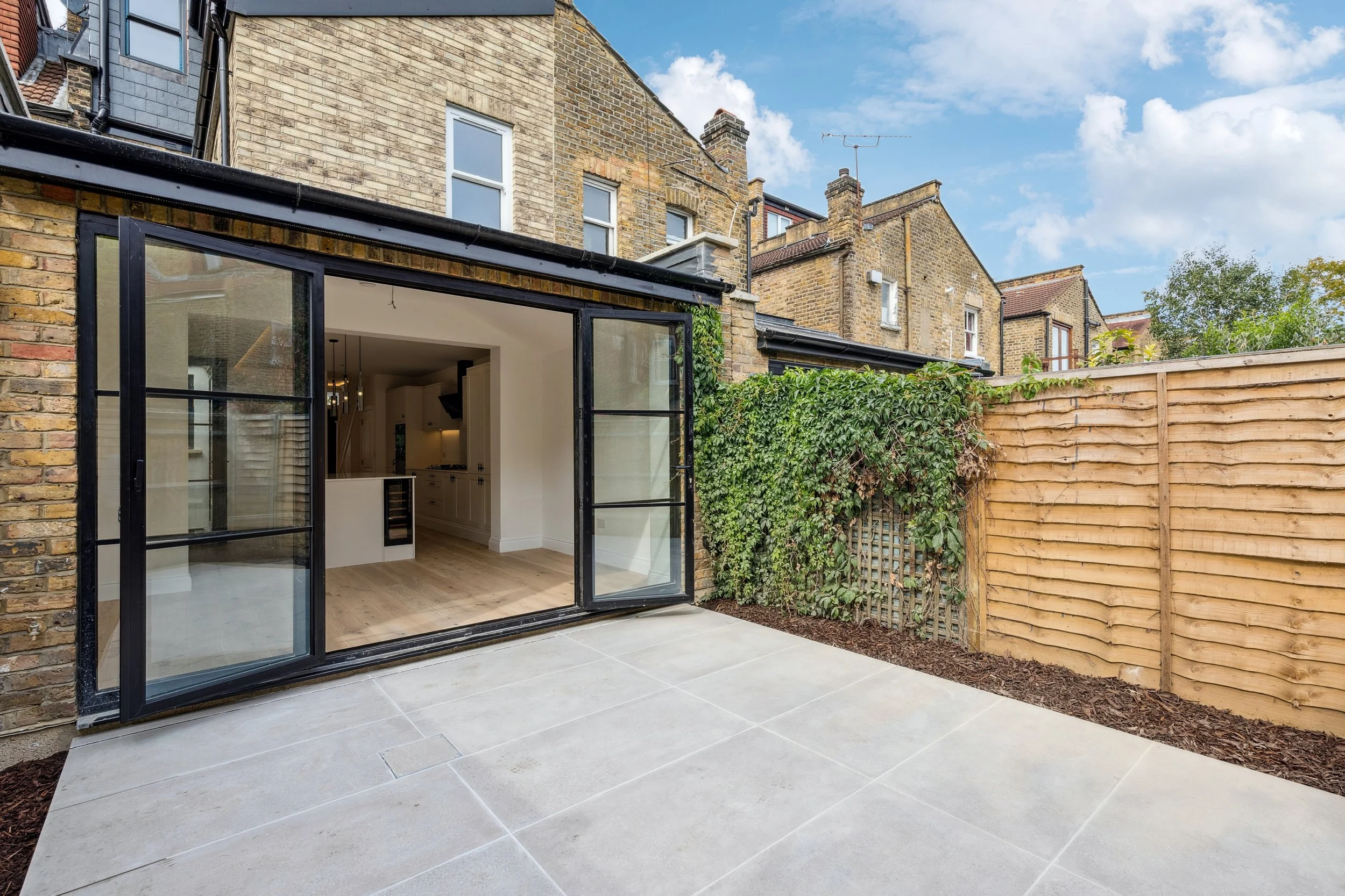 A backyard patio with light grey tiles, an open black-framed glass door leading to a kitchen, a wooden privacy fence, and neighboring brick buildings under a partly cloudy sky with blue sky and white clouds.