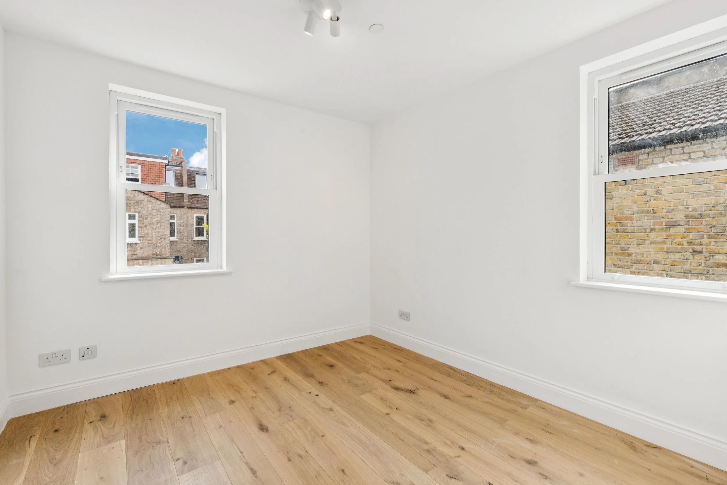 Empty room with white walls, two windows showing neighboring buildings, and wooden flooring.
