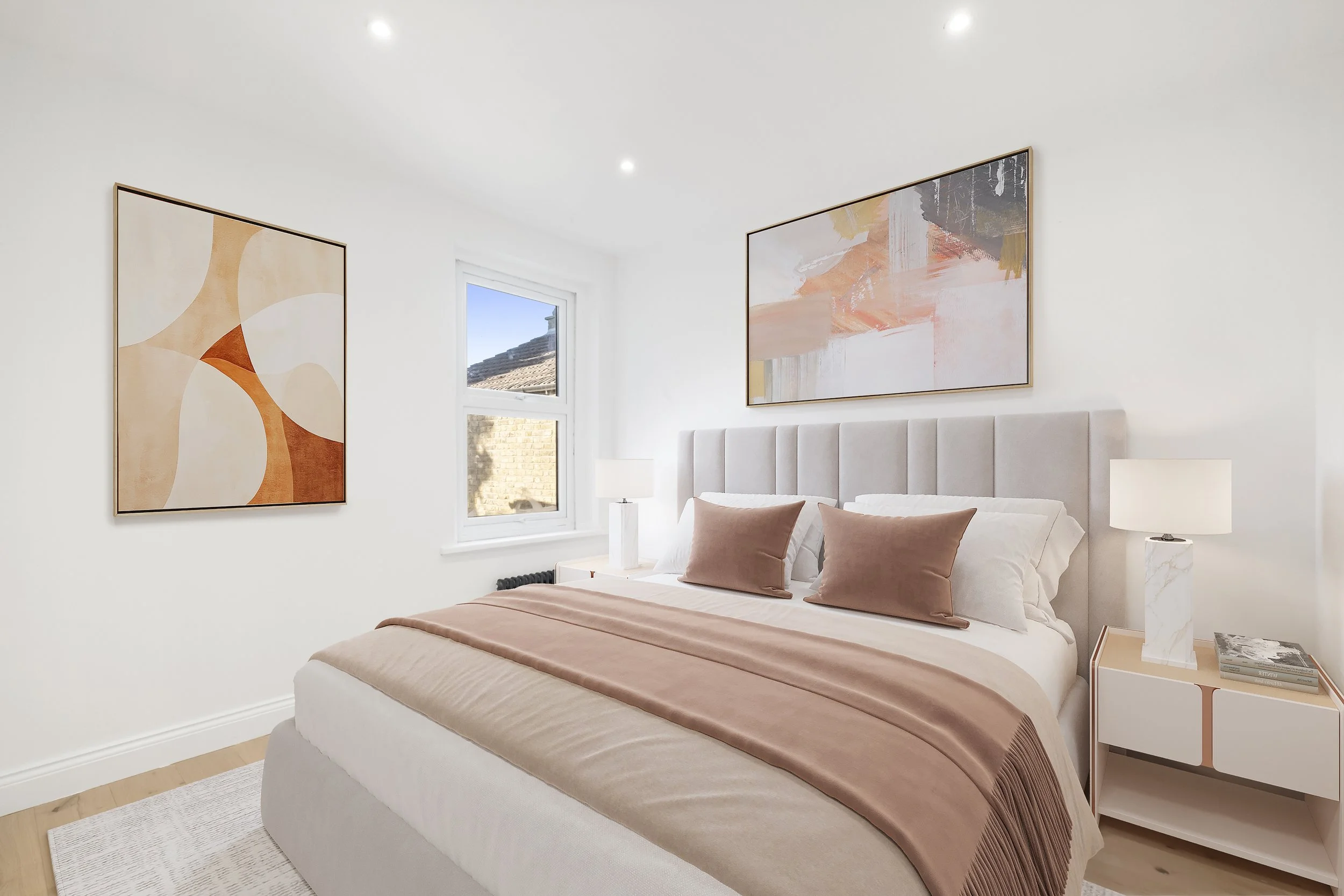 A modern bedroom with a white bed, blush-colored bedspread, matching pillows, two white nightstands, abstract artwork on white walls, a window with a view of a brick wall and roof, and light-colored wooden flooring.