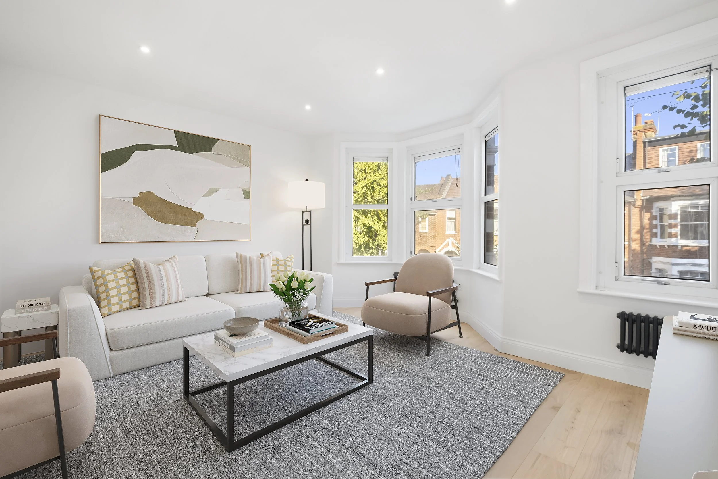 Bright living room with white walls, large windows, a white sofa with patterned pillows, a round beige armchair, a coffee table with books and a flower arrangement, a tall floor lamp, abstract wall art, and a gray area rug.