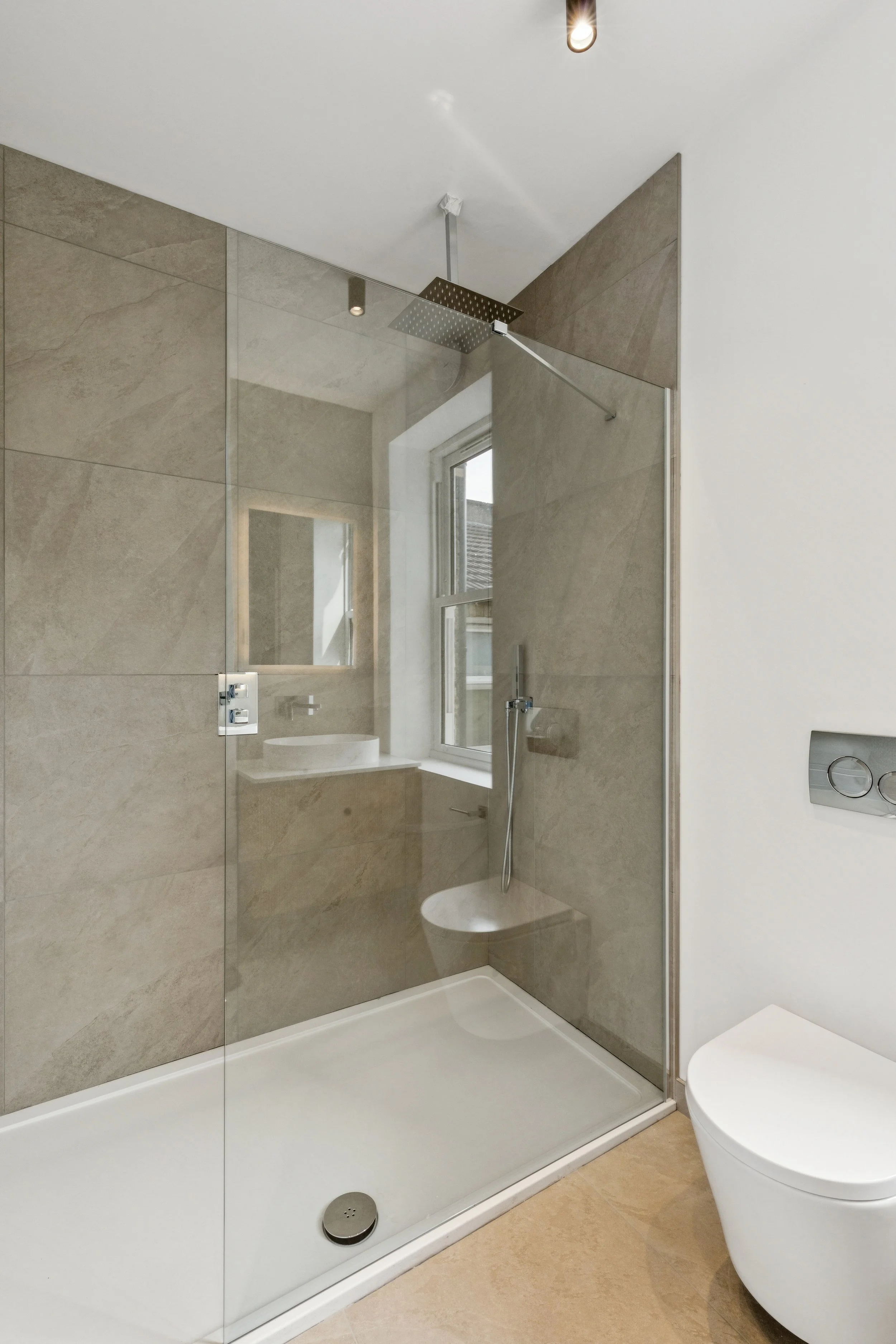 Modern shower area with beige tiled walls, a glass partition, a rain showerhead, a handheld shower, a small built-in seat, a window, and a white toilet nearby.