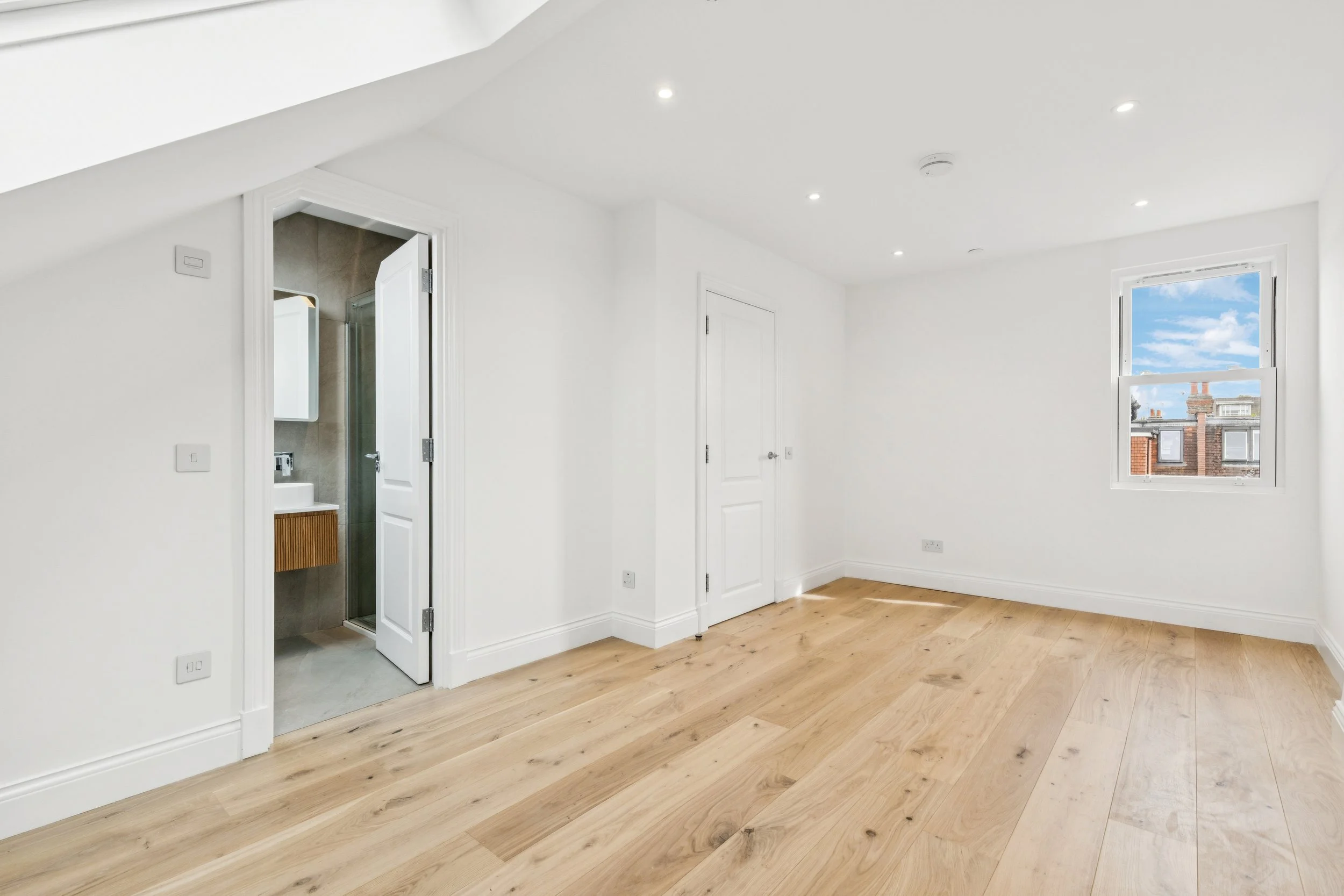 Empty room with wooden flooring, white walls, a window showing a partly cloudy sky and neighboring buildings, and two doors, one leading to a bathroom.