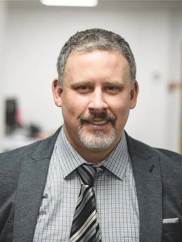 A middle-aged man with short gray hair, a beard, and mustache, dressed in a suit with a checkered shirt and a striped tie, standing indoors with a blurred background.