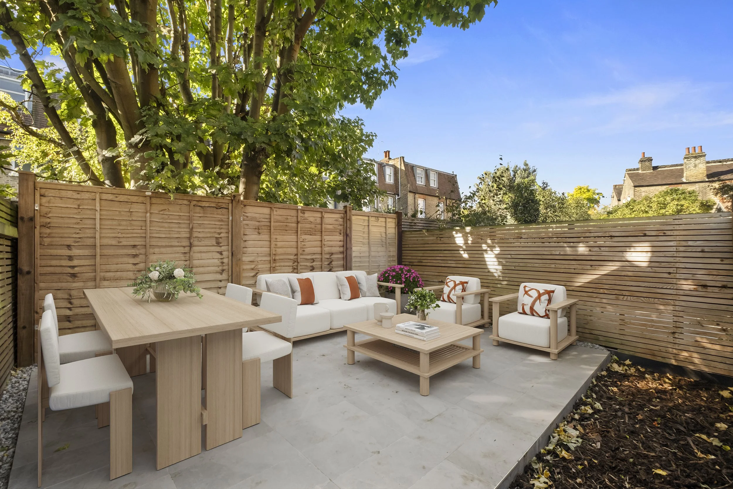 A cozy outdoor patio with a wooden fence, surrounded by a large leafy tree, and neighboring houses visible in the background. The patio features modern white seating with orange and patterned cushions, a wooden dining table with white chairs, and var