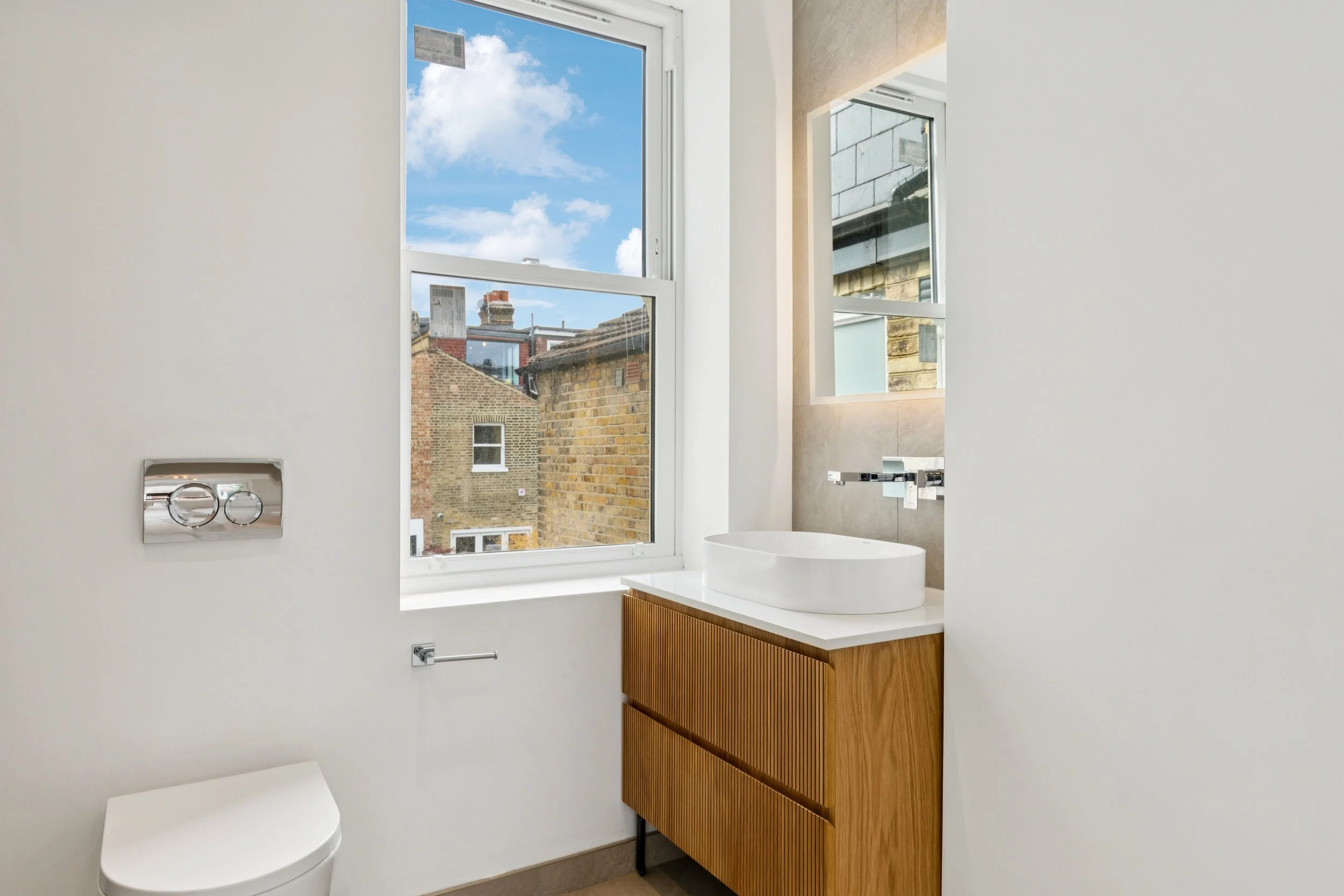 Modern bathroom with large windows showing a blue sky with clouds and neighboring brick buildings. It features a white toilet, a wooden vanity with a white vessel sink, and chrome fixtures.