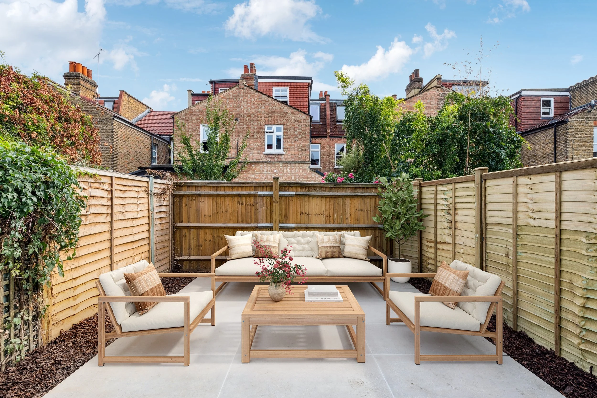 A cozy outdoor seating area with wooden furniture and cream cushions, surrounded by a wooden privacy fence and greenery, with residential buildings and blue sky in the background.
