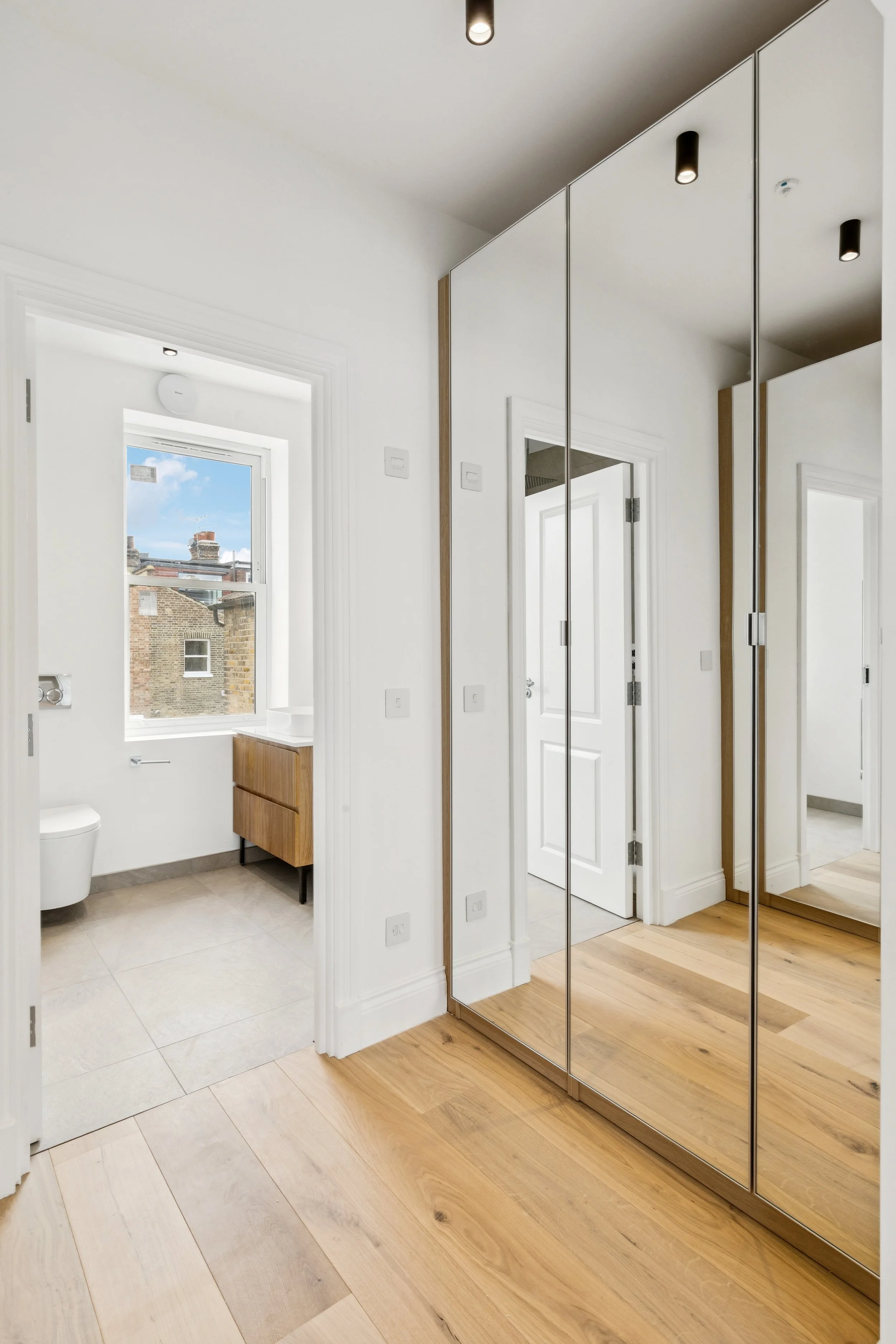 A modern, minimalist bathroom with a large window, a small wooden vanity, and a white toilet. The room has white walls and a tiled floor. A mirrored closet door reflects part of the room, and black ceiling lights are mounted above.