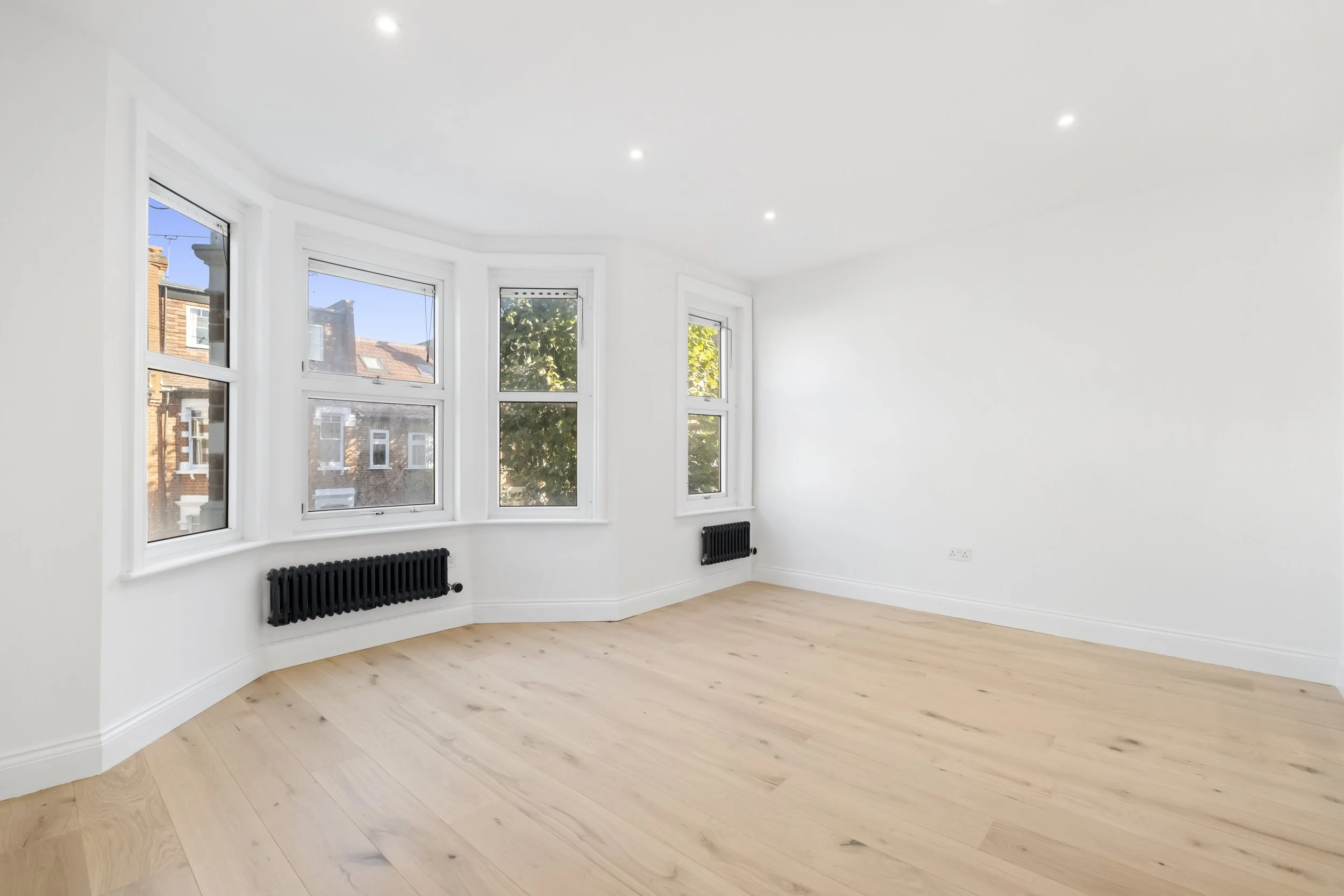 Empty white room with wood flooring, large bay windows, black radiators underneath, and ceiling recessed lights.