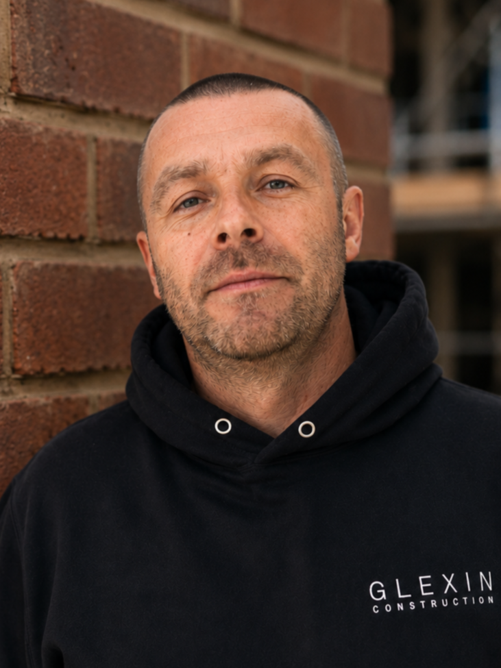 A man with short hair and stubble, wearing a black hoodie with 'GLEXIN CONSTRUCTION' written on it, standing outdoors against a brick wall.