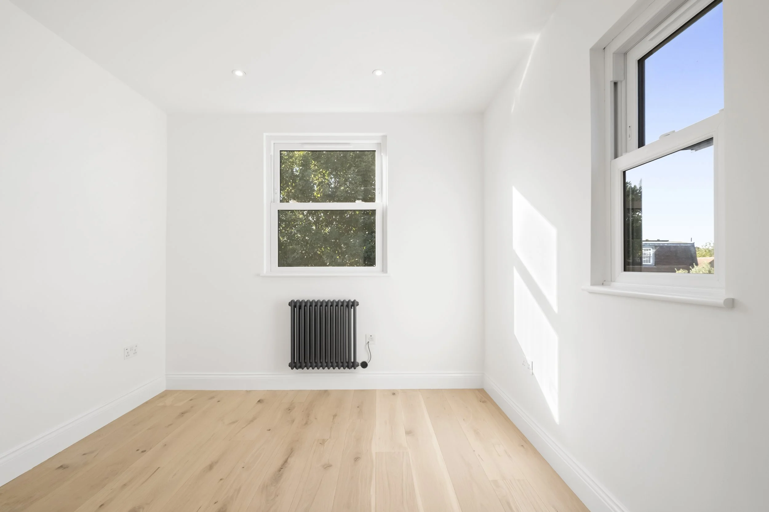 Empty bedroom with white walls, light wood flooring, two white-framed windows, and a black radiator on the wall under the window on the left.