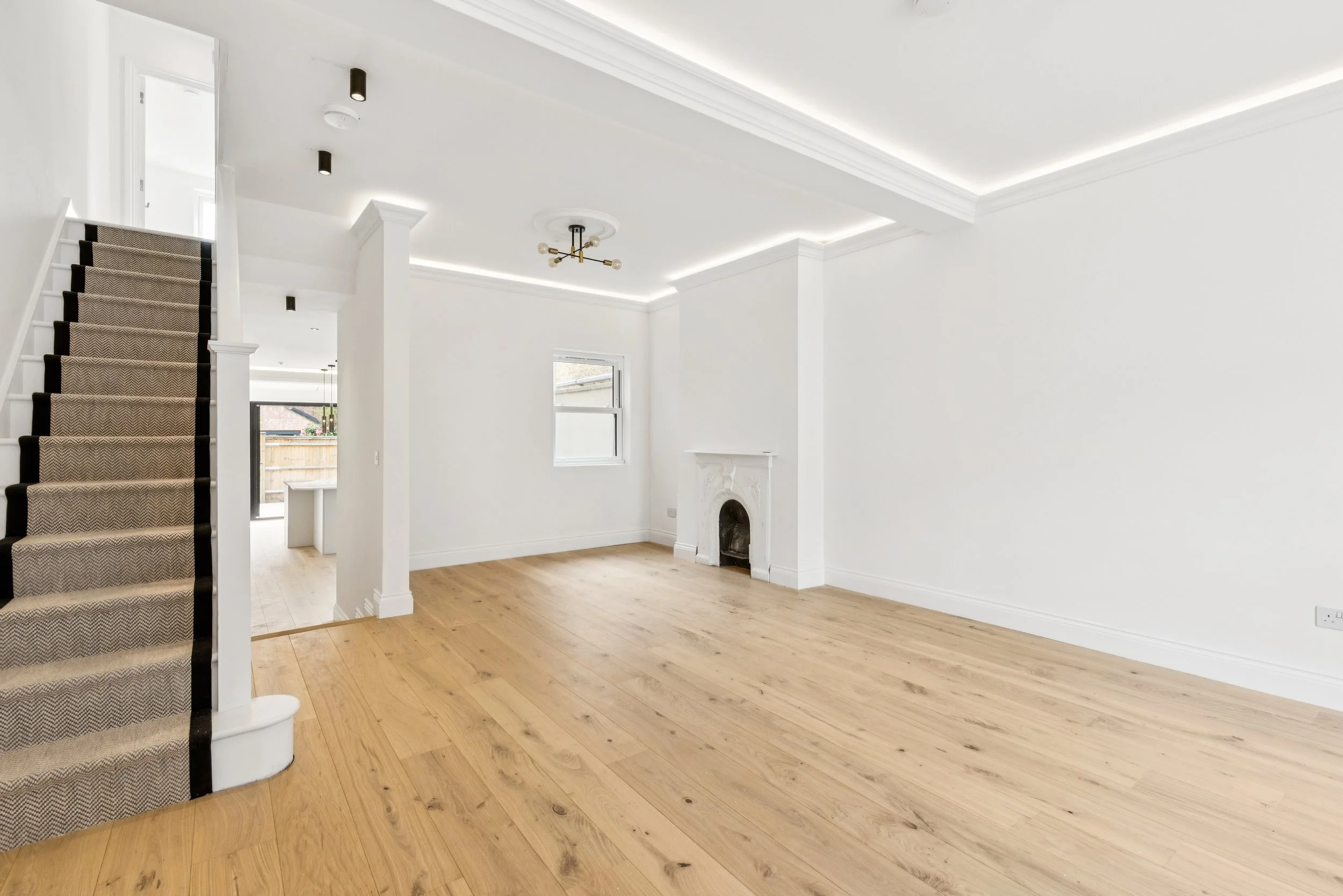 Empty living room with wooden flooring, white walls and ceiling, a fireplace, large windows, and a staircase with beige carpet.