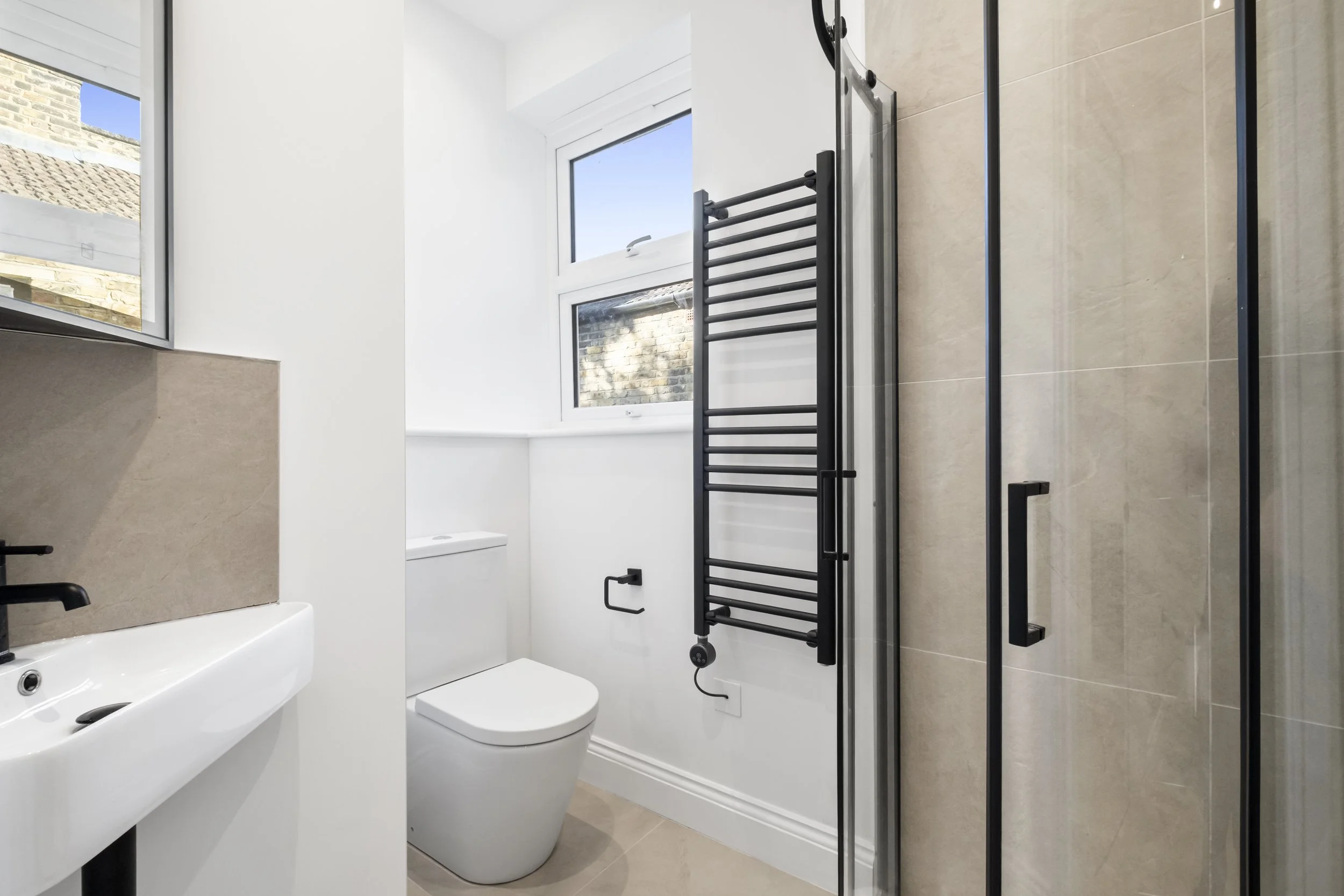 Modern bathroom with white walls, a bathtub, a toilet, a small sink, large windows, beige tiled flooring, a black towel rack, and a glass shower door.