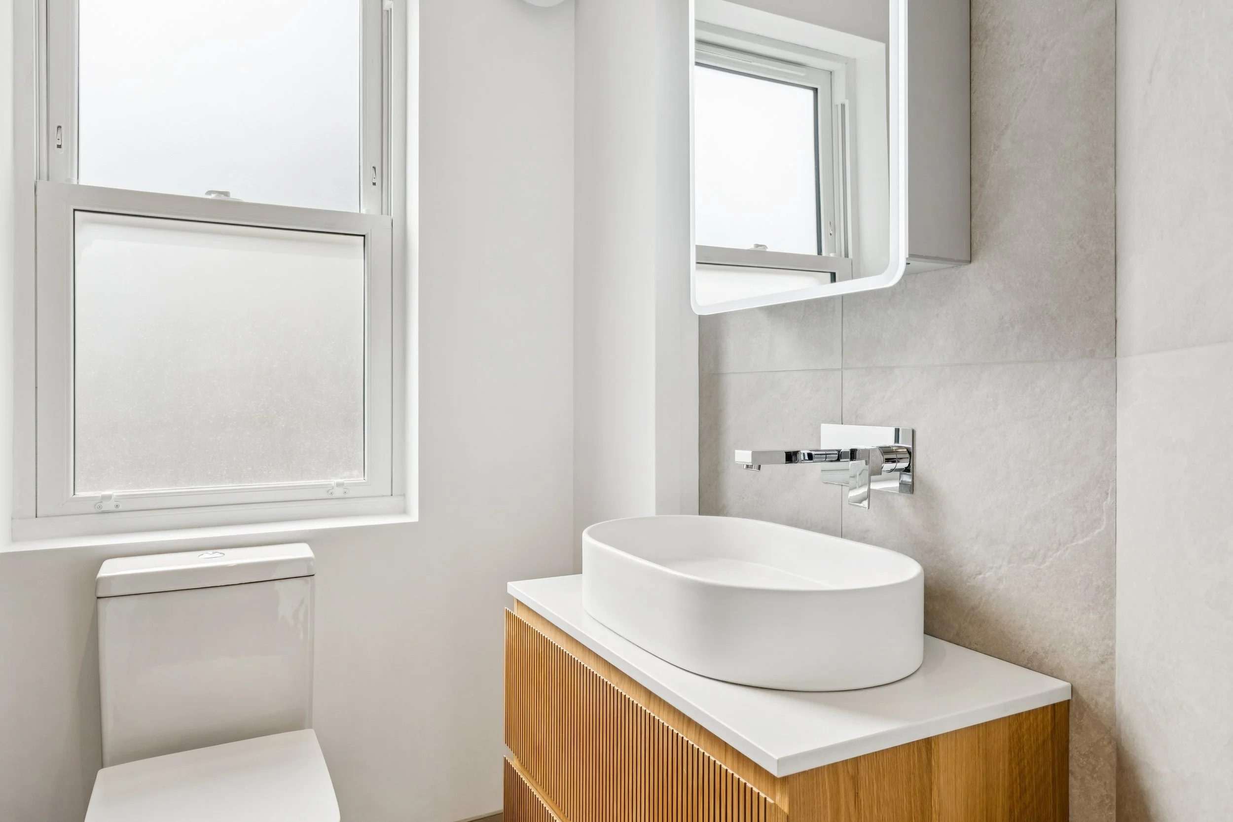Modern bathroom featuring a white vessel sink on a wooden vanity, a wall-mounted faucet, a small mirror, a frosted window, and minimalist decor with neutral tones.
