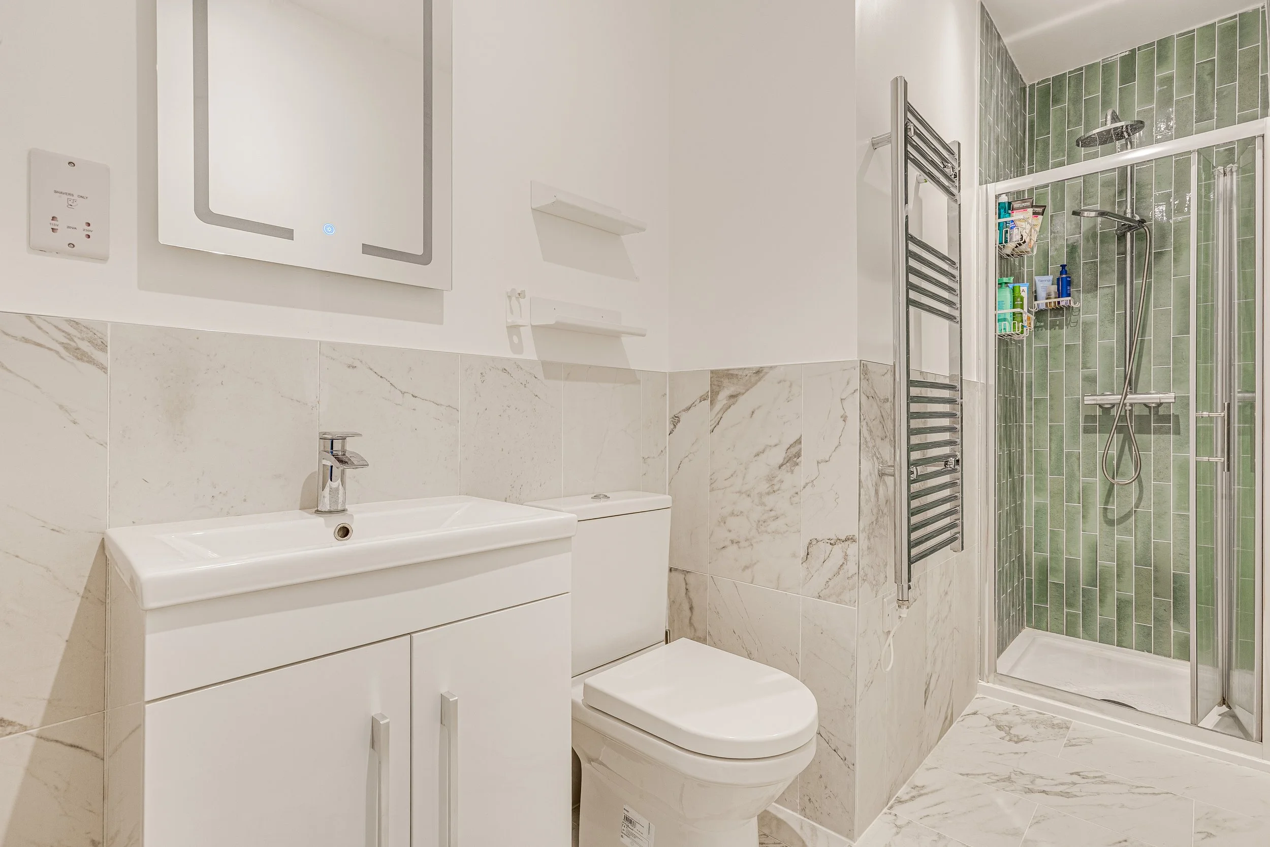 Clean modern bathroom with a white vanity, mirror, toilet, and a glass shower with green tile walls.