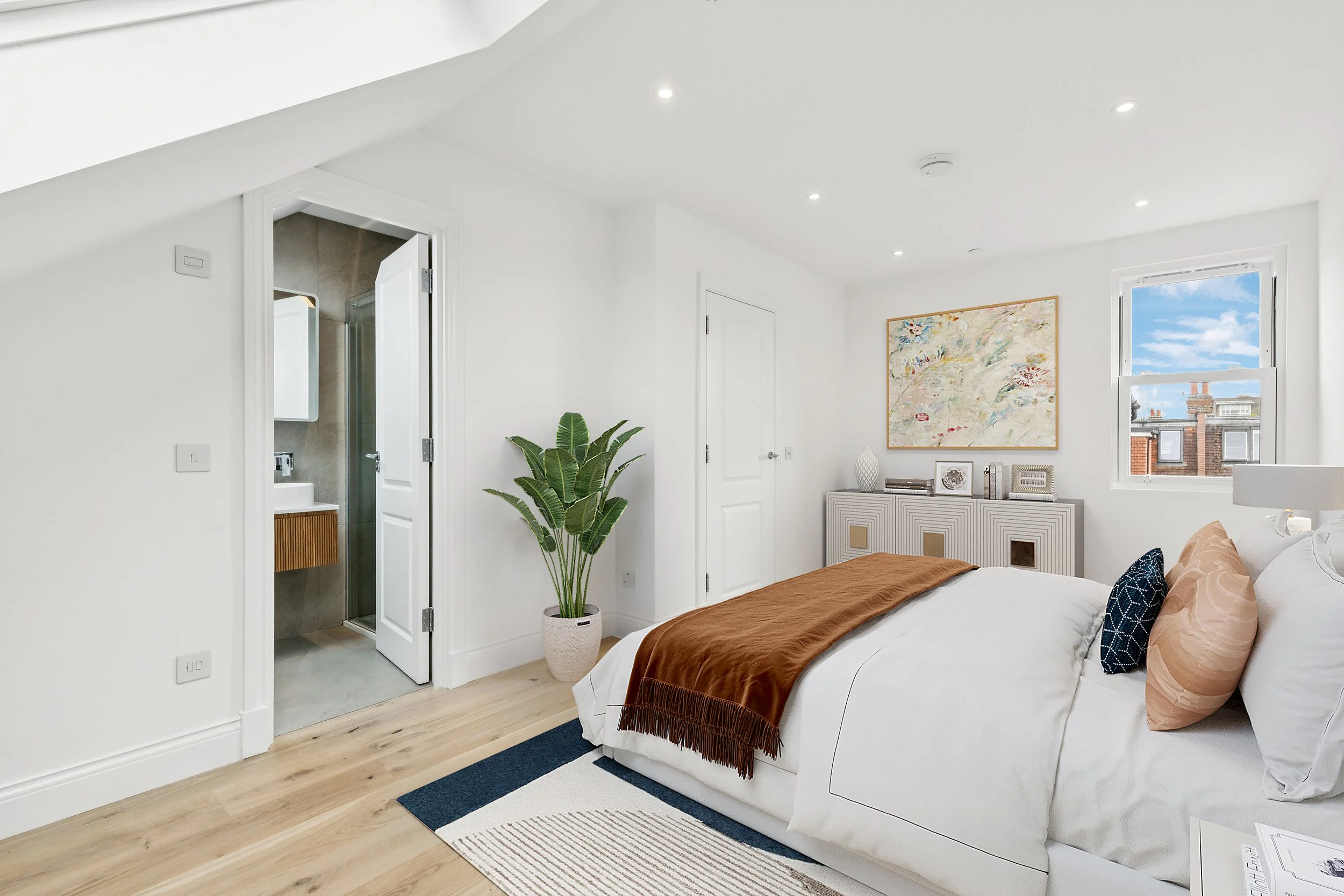 A bedroom with a white bed, brown throw, and decorative pillows, near a window with a cityscape view. There is a large painting above a white console, a potted plant, and a door leading to a bathroom with a shower.