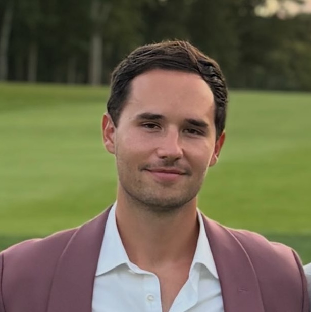 A young man with dark brown hair, wearing a white shirt and a maroon blazer, standing outdoors on a grassy area with trees in the background.