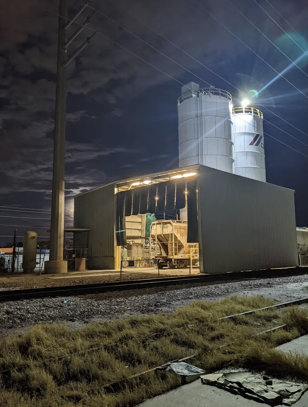 A night view of an industrial building with large metal silos and a train car parked inside a covered area, with a utility pole and power lines overhead, and a cloudy sky in the background.