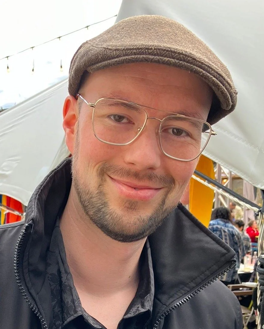 A young man smiling outdoors, wearing glasses, a brown cap, and a black jacket, at what appears to be a festive event or market.