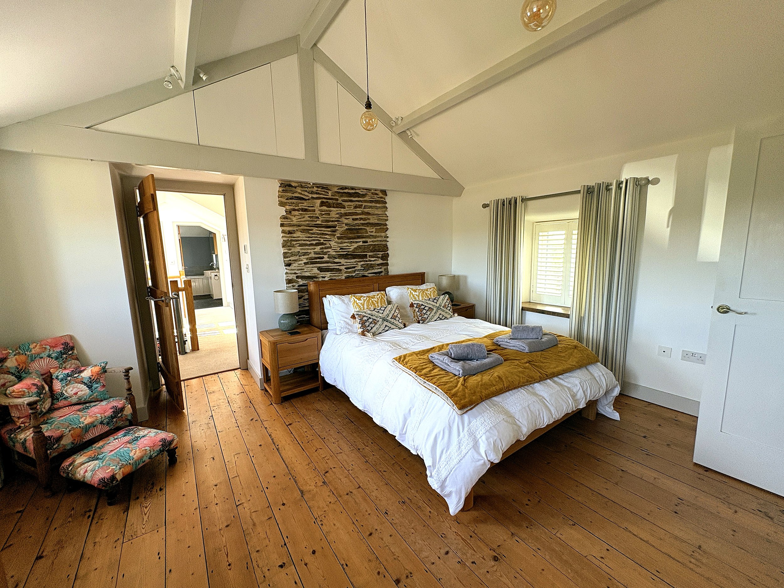 A bedroom with a wooden floor, a large bed with white and mustard-colored bedding, a stone accent wall behind the bed, and windows with striped curtains. There is a floral armchair on the left and a partially opened door leading to a bathroom.