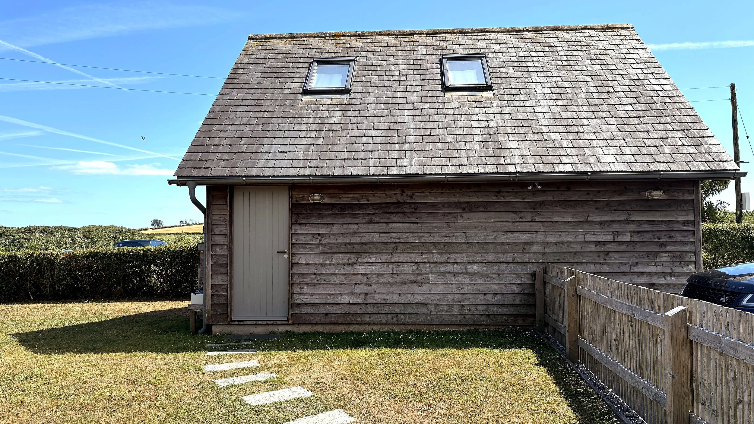 A wooden house with a steep, shingled roof and two small skylight windows against a blue sky. The house has a white door, a fenced yard with stepping stones, and a backup vehicle partially visible.
