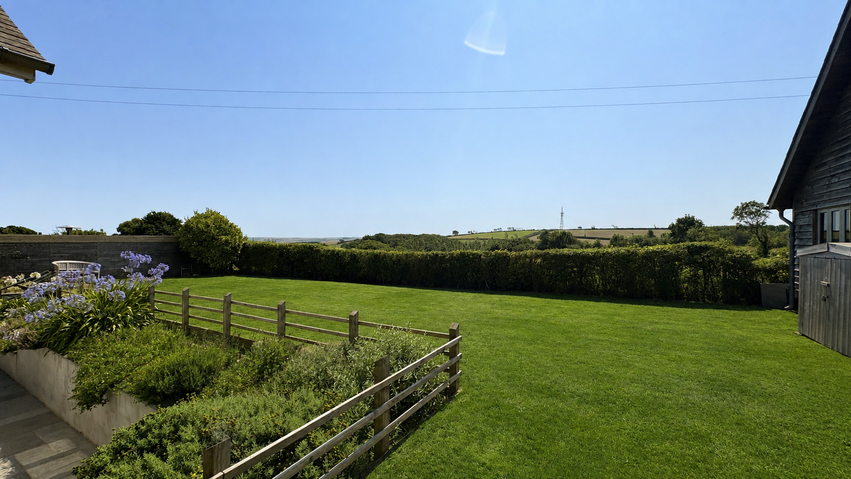 A backyard with a green lawn, wooden fence, flowering plants, and a large tree, with rolling hills and blue sky in the background.