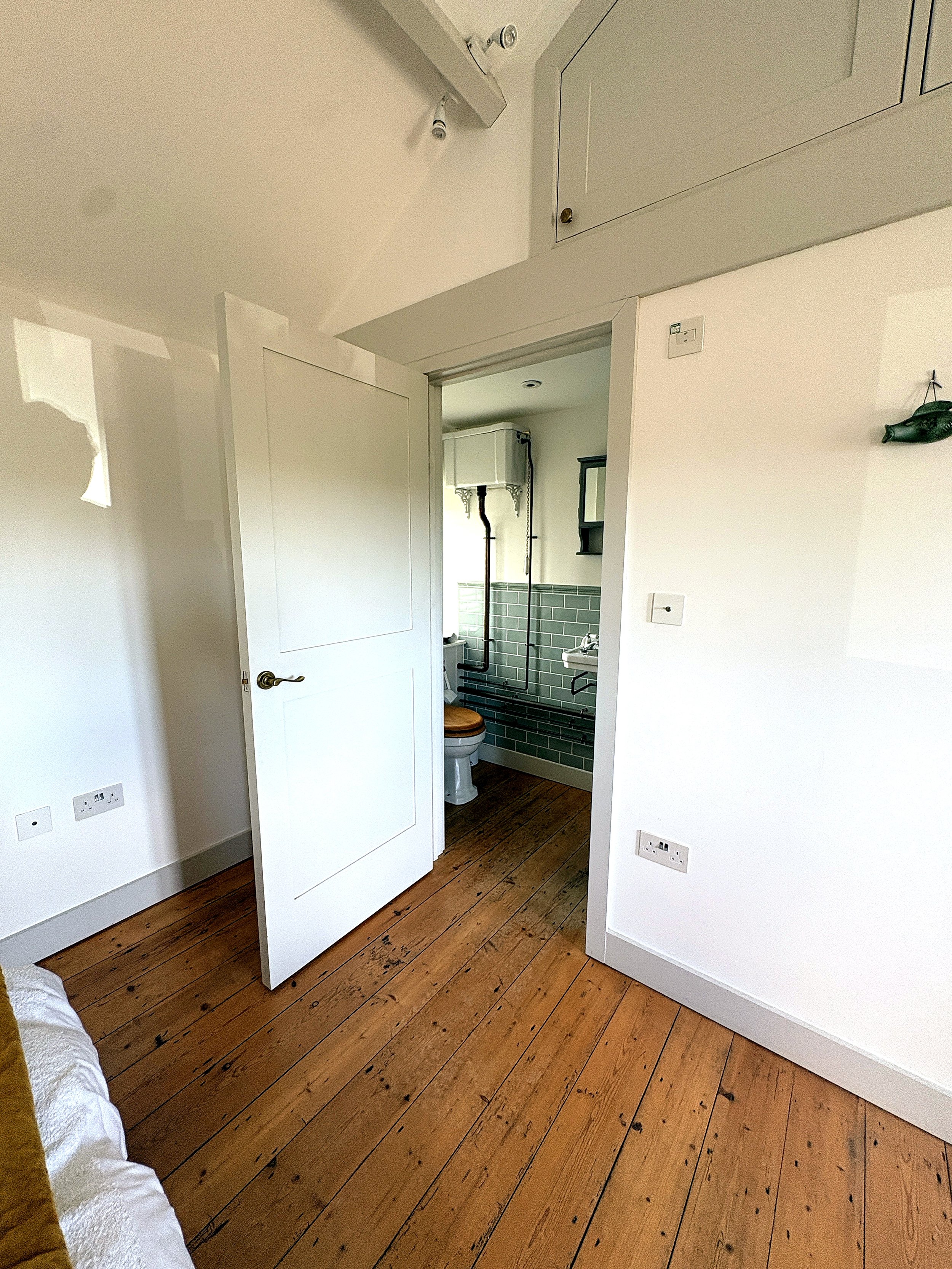 A partially open white door leading into a small bathroom with a toilet and green tiled wall. The room has white walls, wooden floorboards, and visible electrical outlets.
