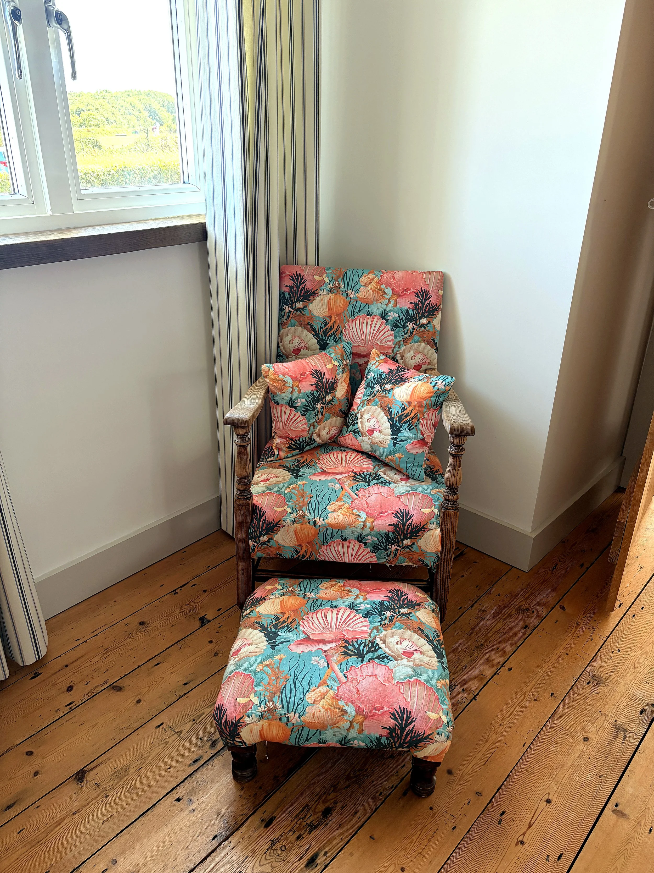 A wooden armchair with a floral cushion and matching pillow in a corner near a window with striped curtains and a view of green trees outside.