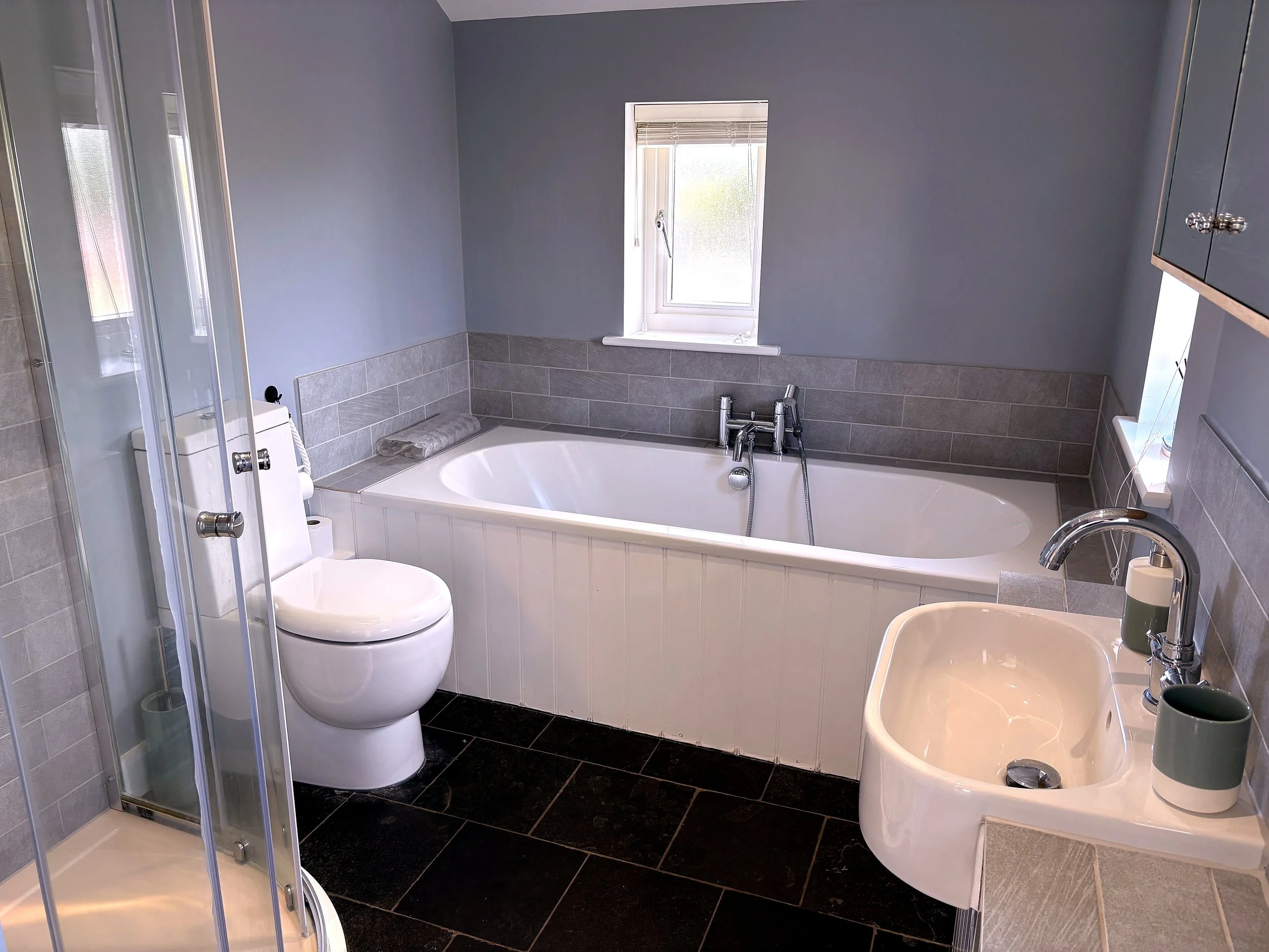 A modern bathroom featuring a corner bathtub with chrome fixtures, a small window with blinds, a toilet with a bidet attachment, a clear glass shower enclosure, a sink with a soap dispenser, and dark tiled flooring.
