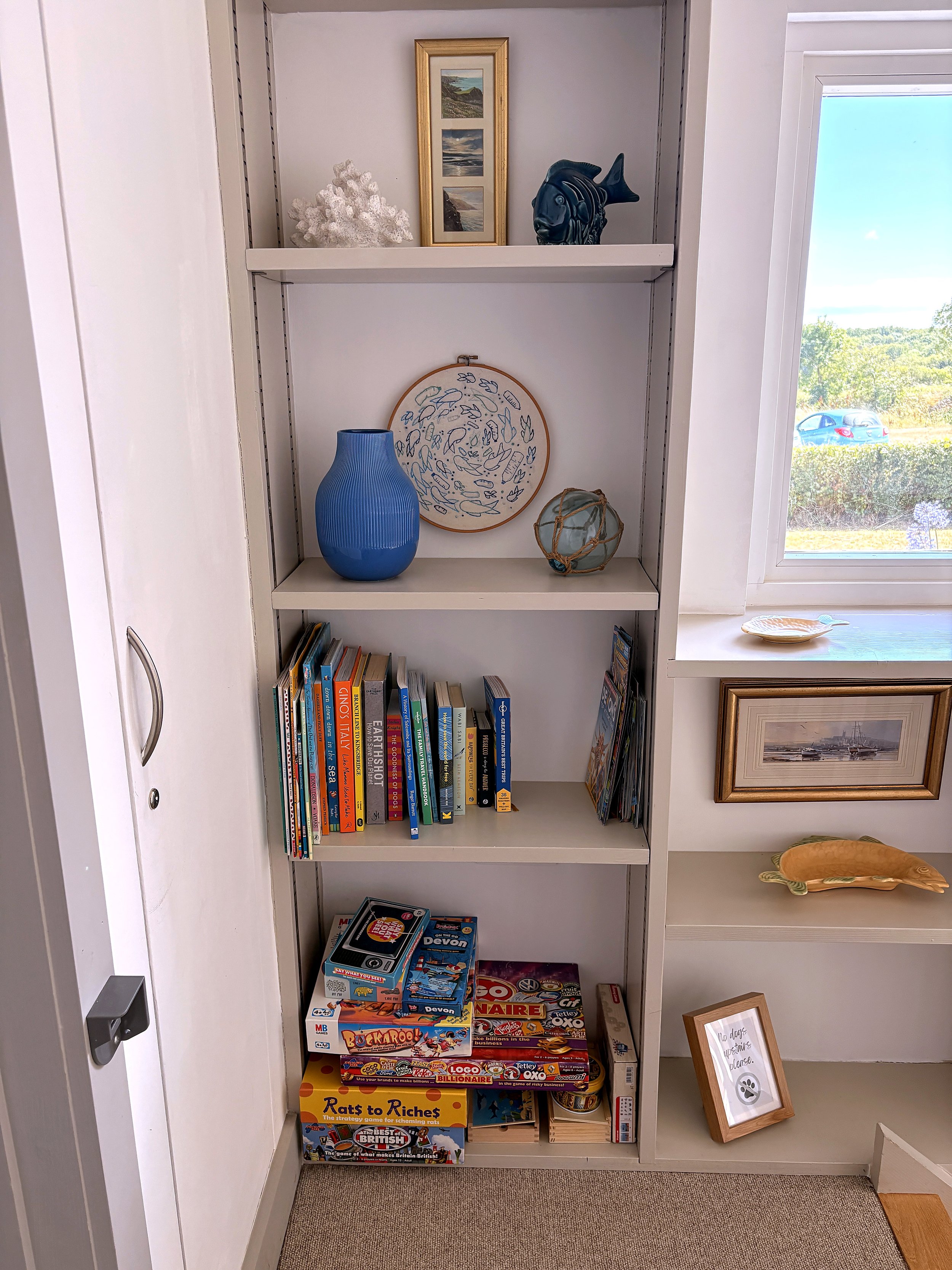 A white bookshelf with decorative items on the top shelf, including a framed picture, a piece of white coral, and a black fish sculpture. The middle shelf holds a blue vase and a round embroidery hoop with a leaf pattern. The bottom shelf contains bo