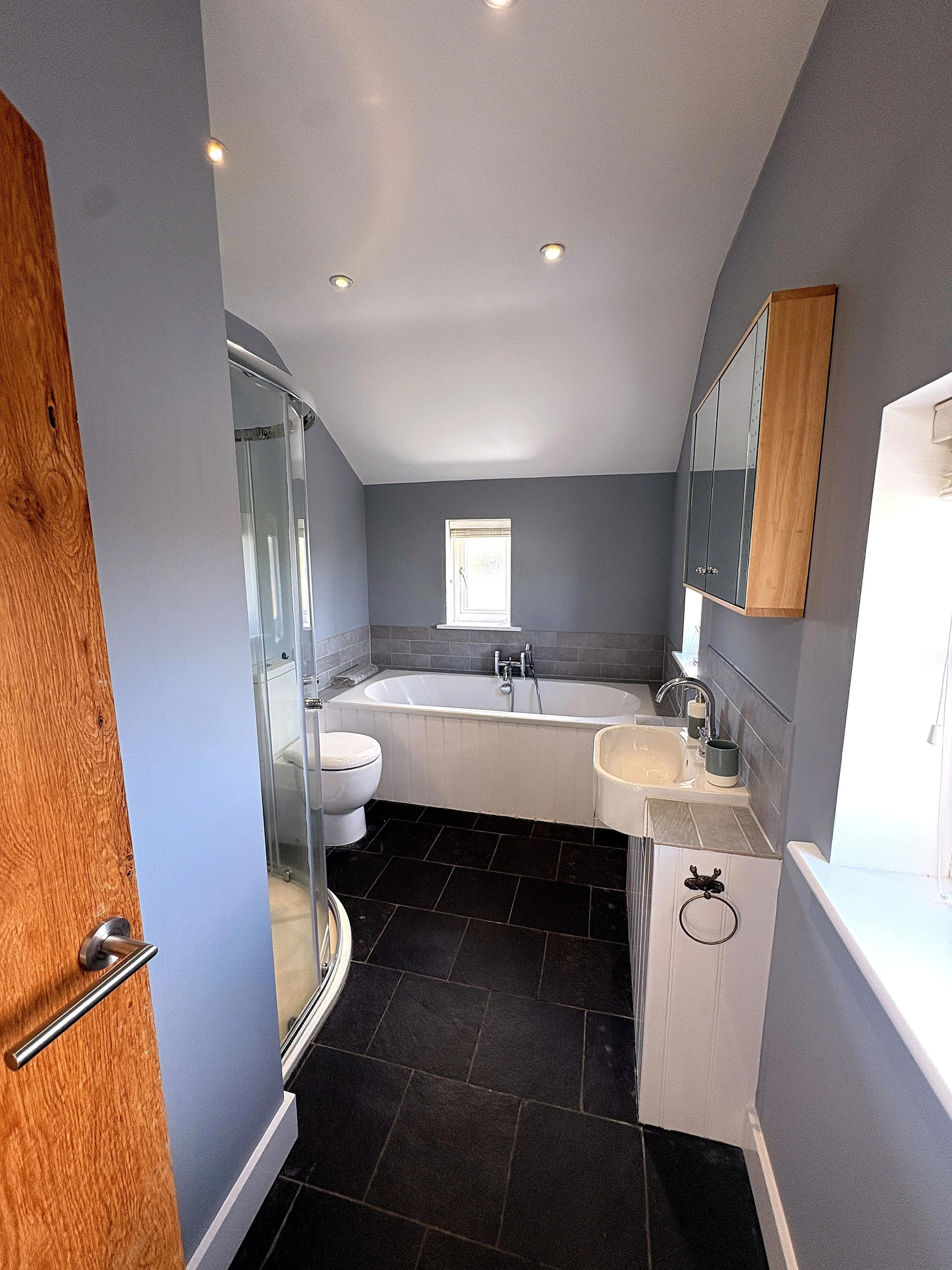 A bathroom with a corner bathtub, a toilet, a small window, a sink, a mirror cabinet, grey walls, black tile flooring, and a shower enclosure.
