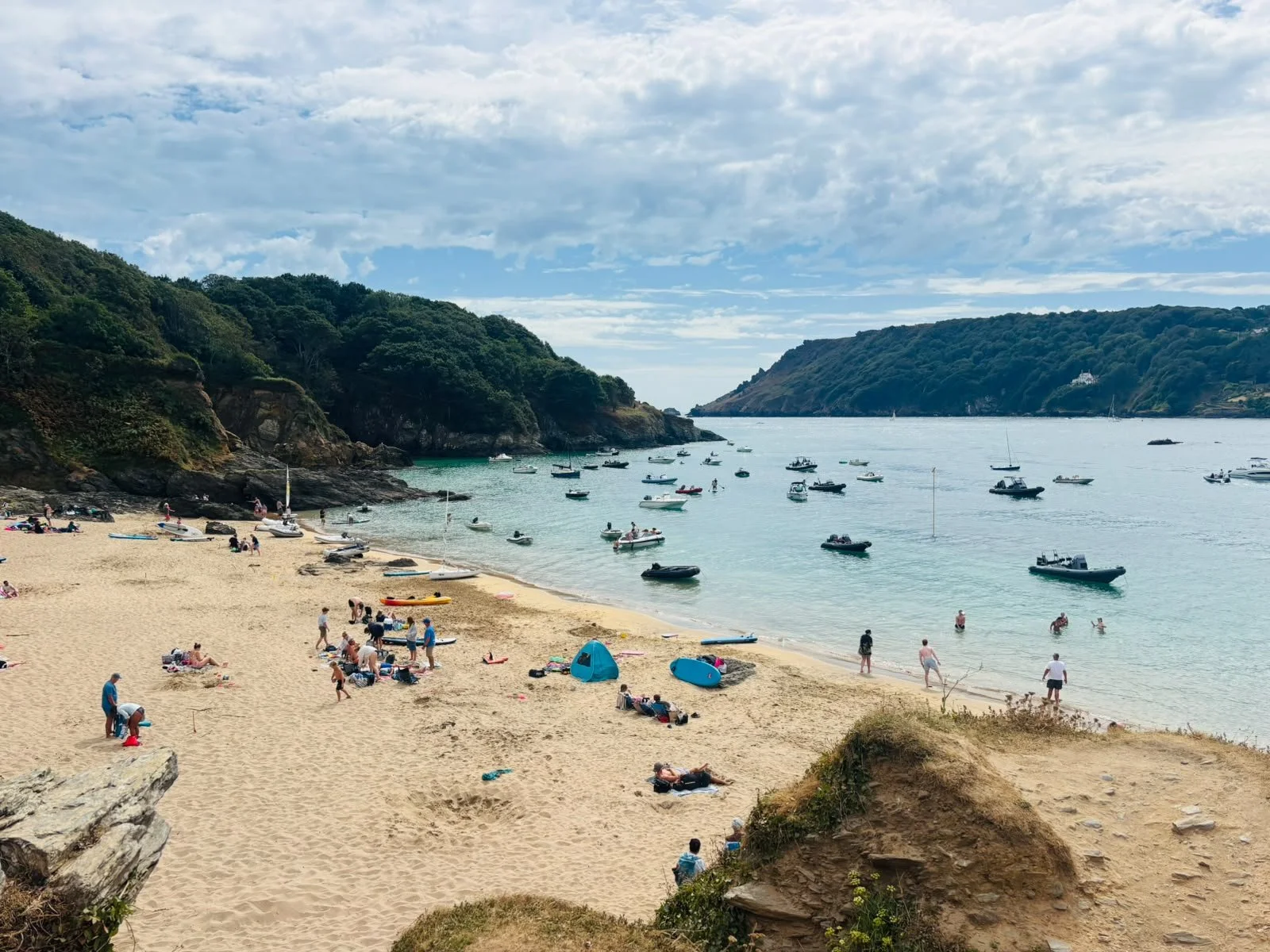 A crowded beach with people sunbathing and swimming, boats anchored in the water, and green hills in the background under a partly cloudy sky.