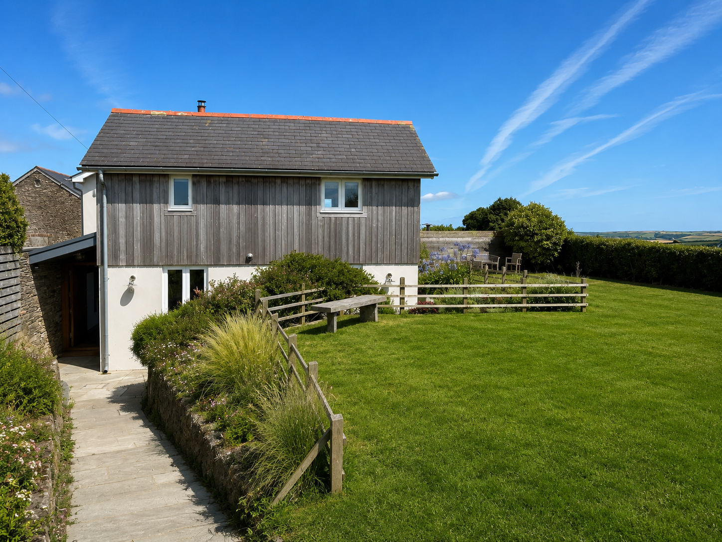 A two-story house with gray wooden siding and a red-tiled roof, surrounded by a green lawn and garden, under a blue sky with wispy clouds.