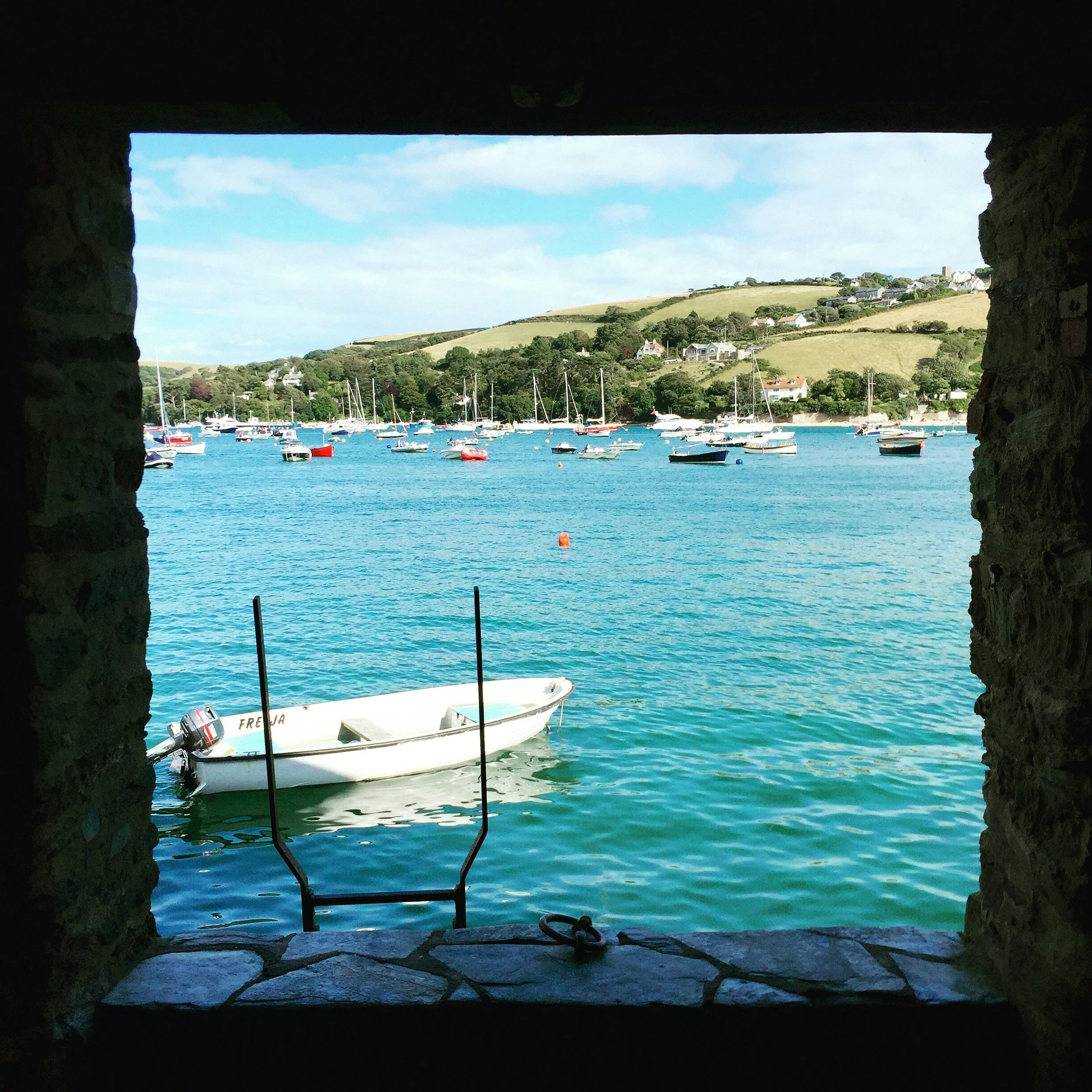 View through a stone-framed window showing boats anchored on a harbor with a hillside village and blue sky in the background.