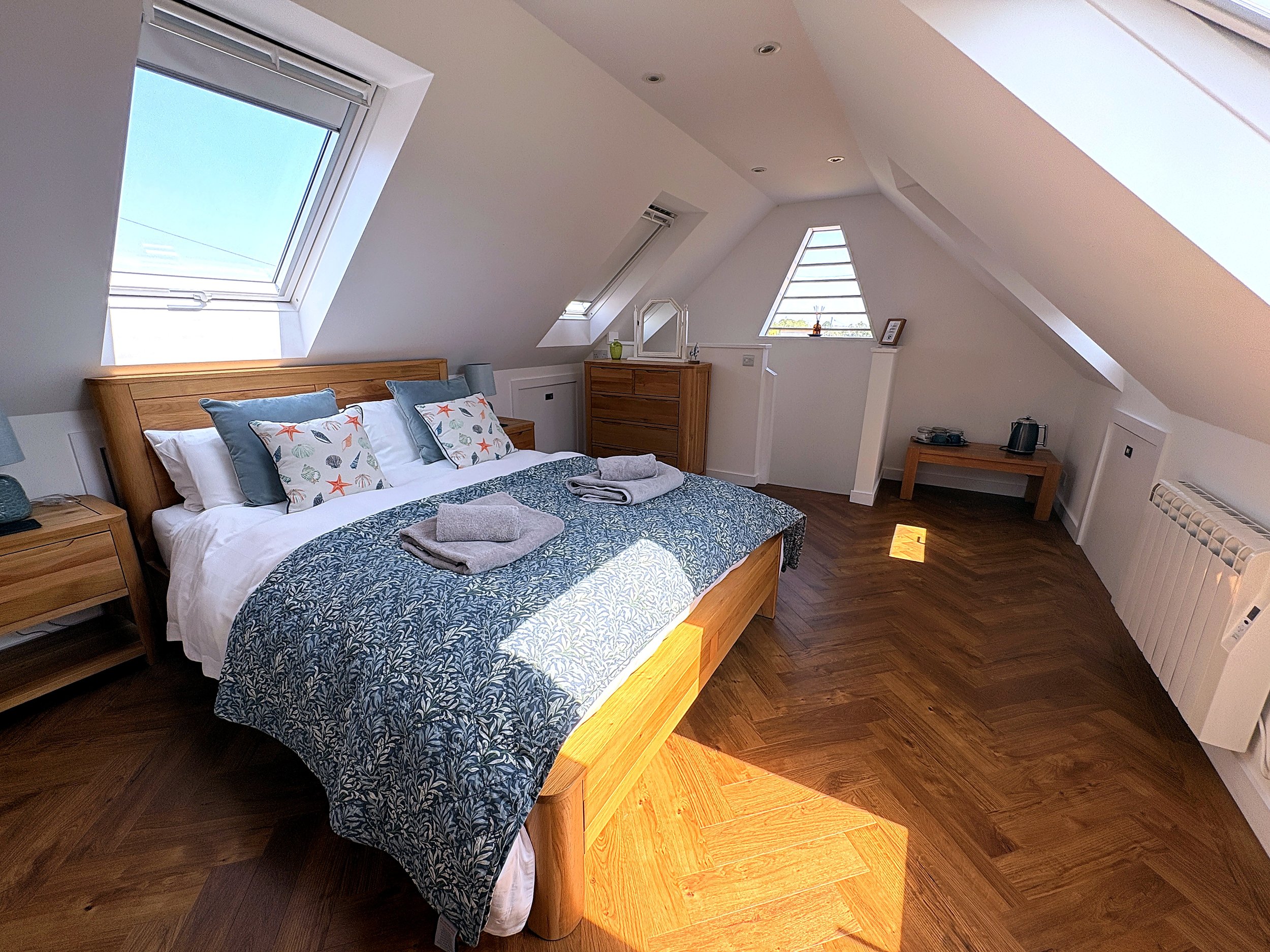 A bedroom with a wooden bed, decorated with pillows and a bedspread, under multiple skylight windows on slanted ceilings, wooden flooring, and furniture including a dresser with mirror, a small side table with a kettle, and a framed picture.