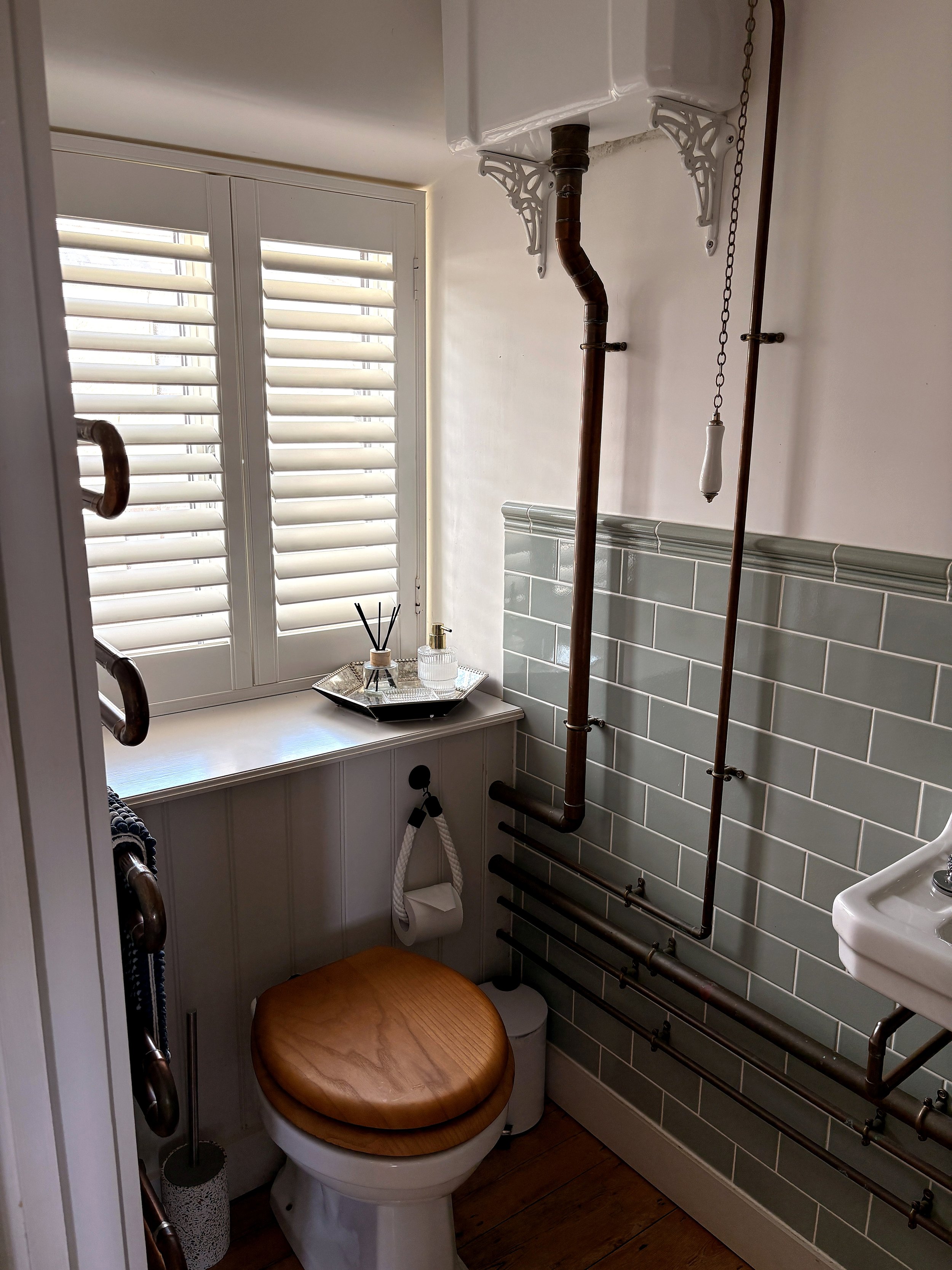 A small bathroom with a toilet with a wooden seat. The wall behind the toilet has ceramic tiles, and there are copper pipes running vertically on the wall. A window with white plantation shutters lets in natural light. On a small ledge above the toil