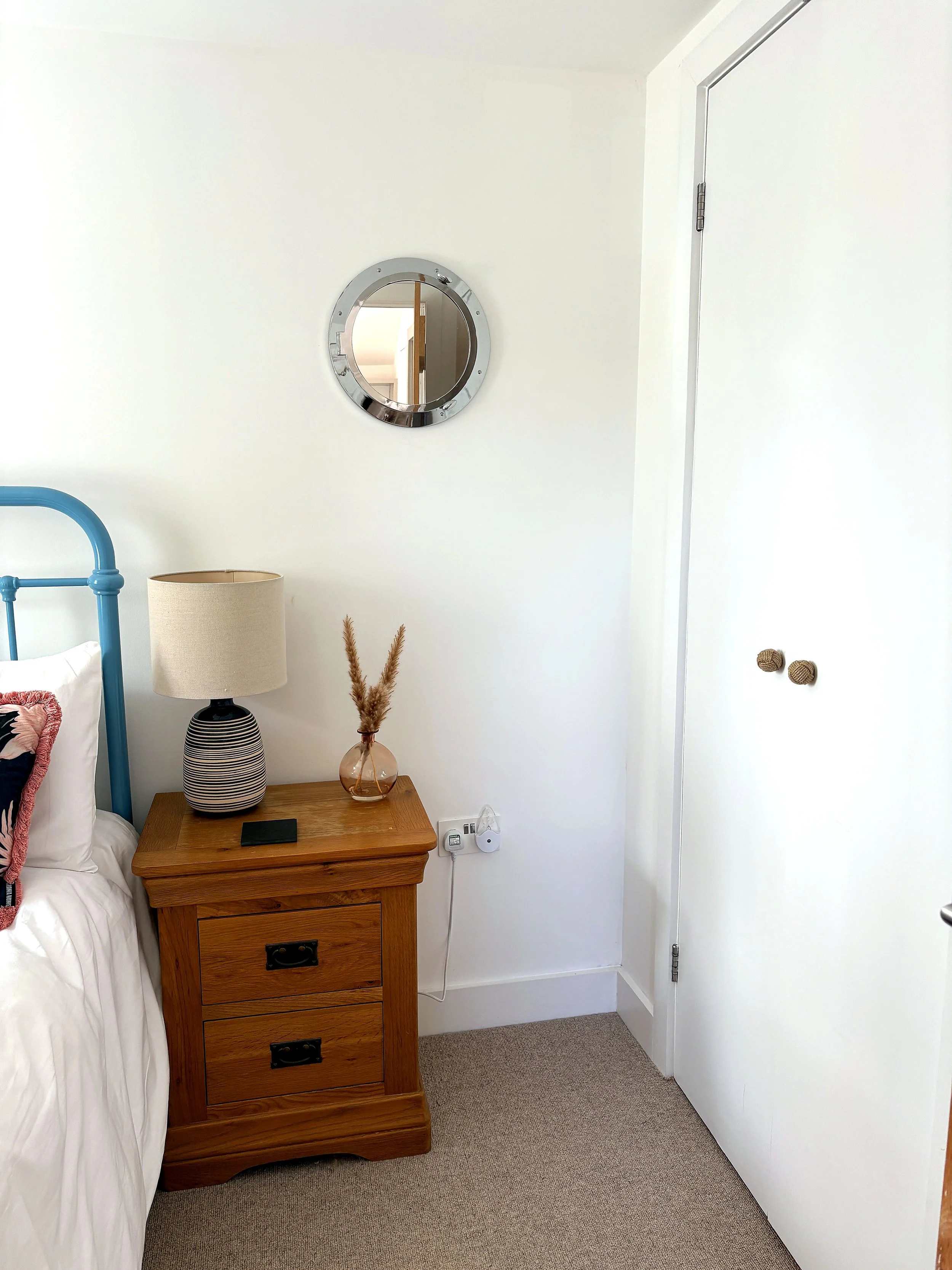 A bedroom corner with a wooden nightstand, a striped table lamp, a small glass vase with dried plants, a blue bed frame, part of a bed with white bedding, and a mirror on a white wall above the nightstand. A door with round knobs is also visible.