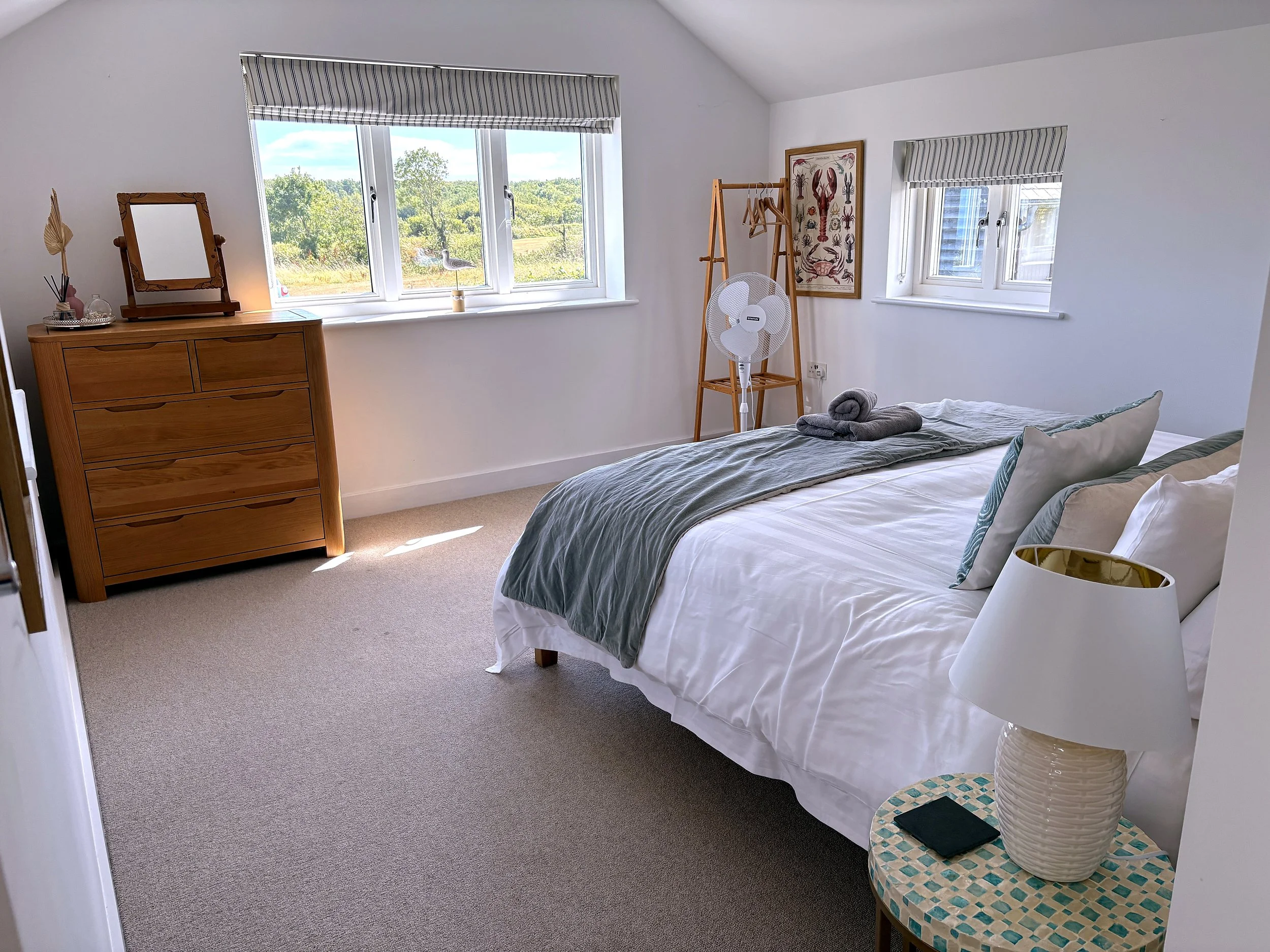 A bedroom with a bed covered in white sheets and a gray blanket, two pillows, and towels folded on top. There are two small windows with striped gray and white shades, a wooden dresser with a mirror and decor items, a fan, and a wooden clothing rack.
