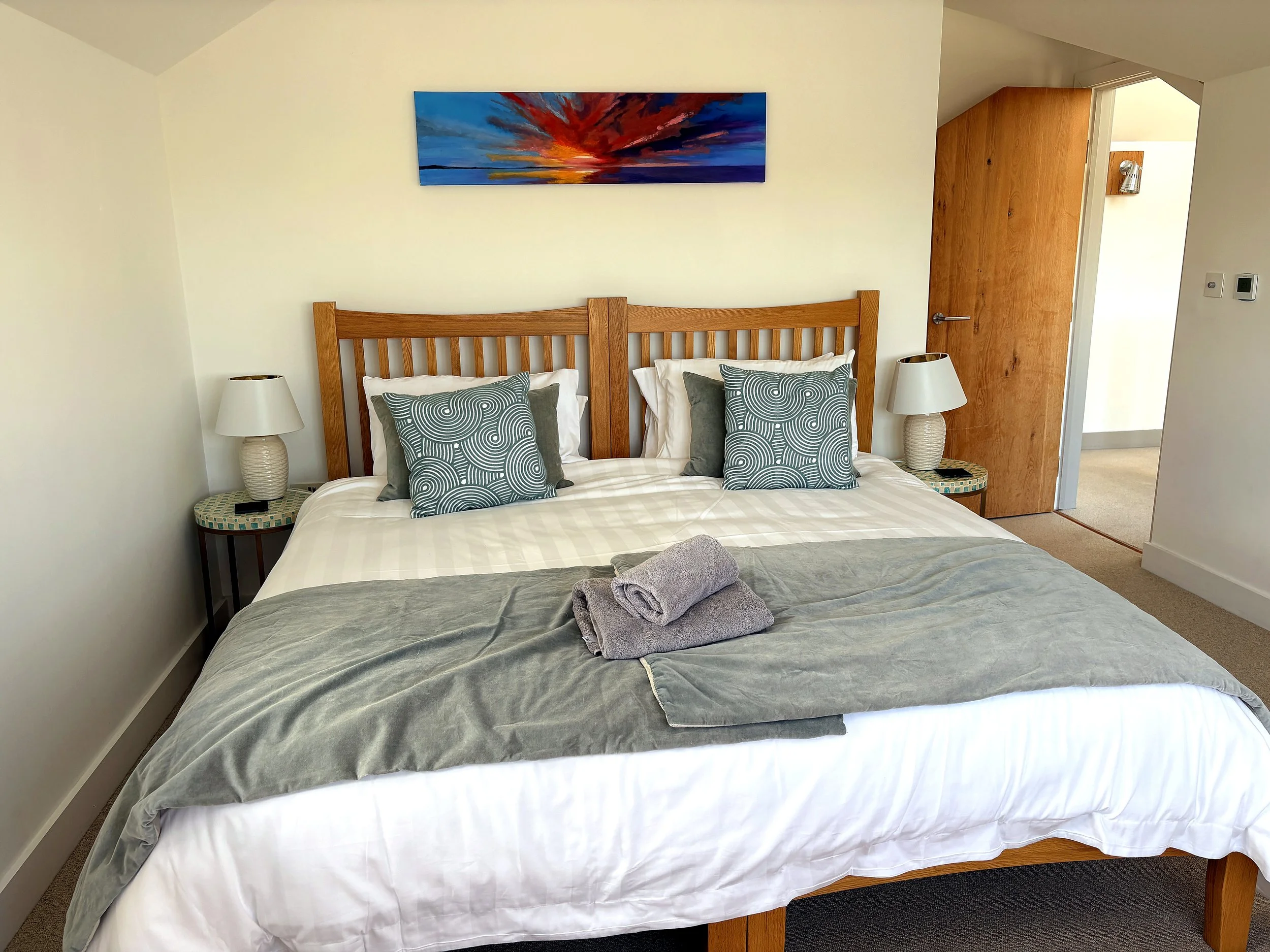 A neatly made bed in a bedroom with a wooden headboard, two decorative pillows with swirl patterns, a white comforter, and a gray blanket. Two matching bedside lamps are on round side tables on each side of the bed. A framed colorful sunset painting 