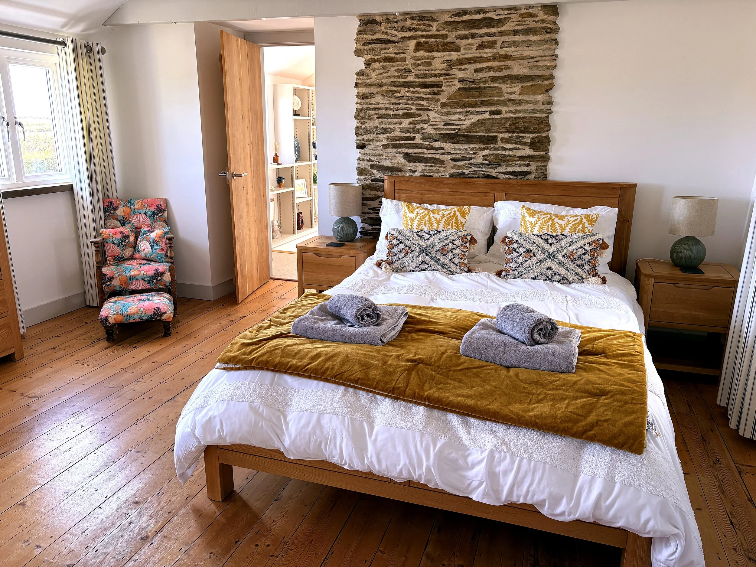 A bedroom with a wooden bed frame, white bedding with yellow and patterned pillows, two bedside tables with lamps, a colorful patterned armchair, a stone accent wall, and an open doorway leading to a room with shelves.