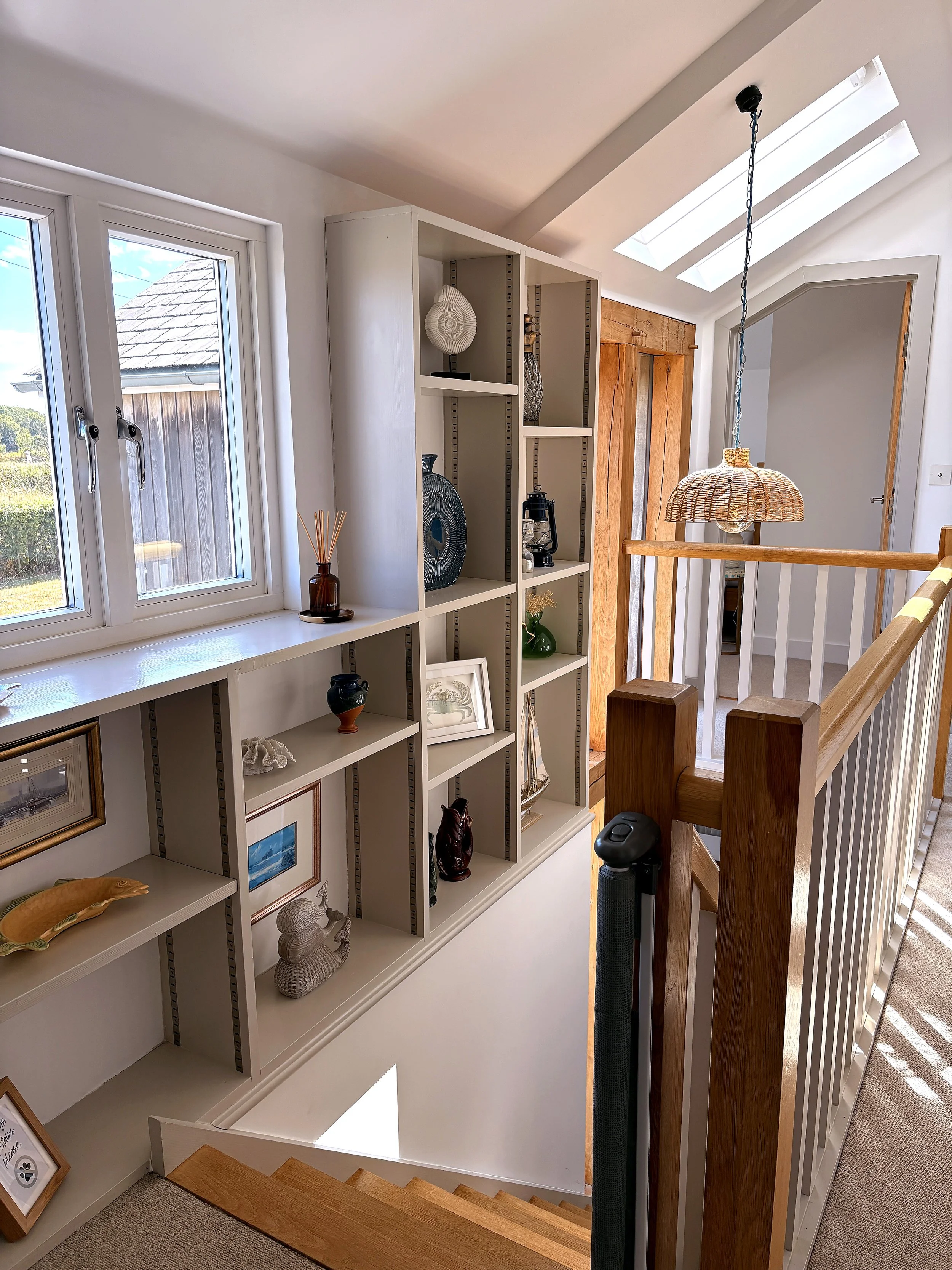 Interior view of a landing at the top of a staircase with a white bookshelf filled with decorative items, a window, a wooden door, and skylights in the ceiling.