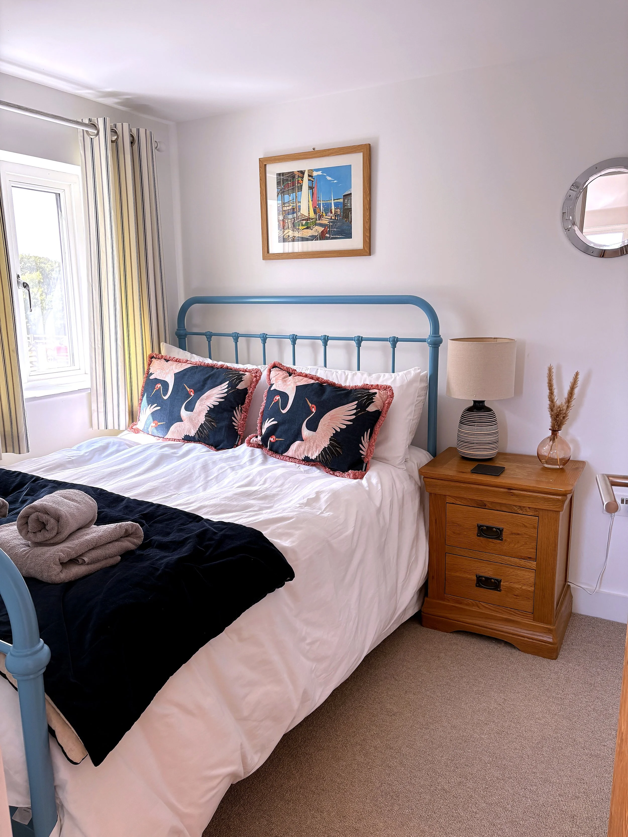 A bedroom with a bed having decorative pillows, a bedside table with a lamp and decorative vase, a picture on the wall, and a window with striped curtains.