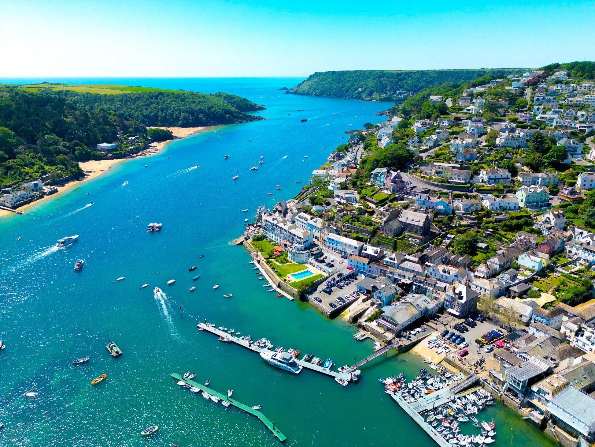 Aerial view of a coastal town with colorful houses along the shoreline, a marina with boats, a beach, and green hills in the background on a sunny day with clear blue sky.