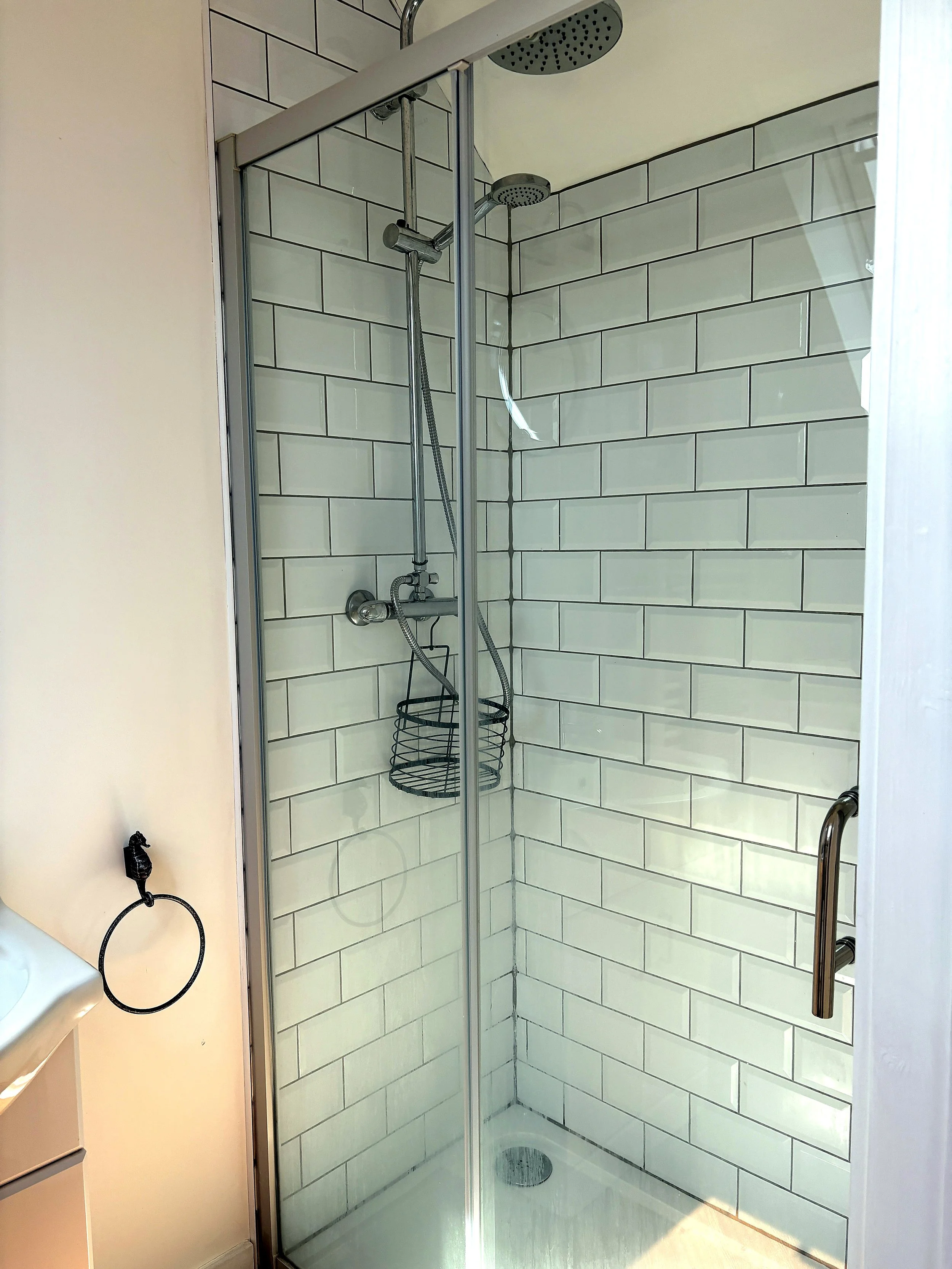 Inside a corner shower with glass door, white subway tile walls, silver showerhead and fixtures, and a wire shower caddy hanging inside.