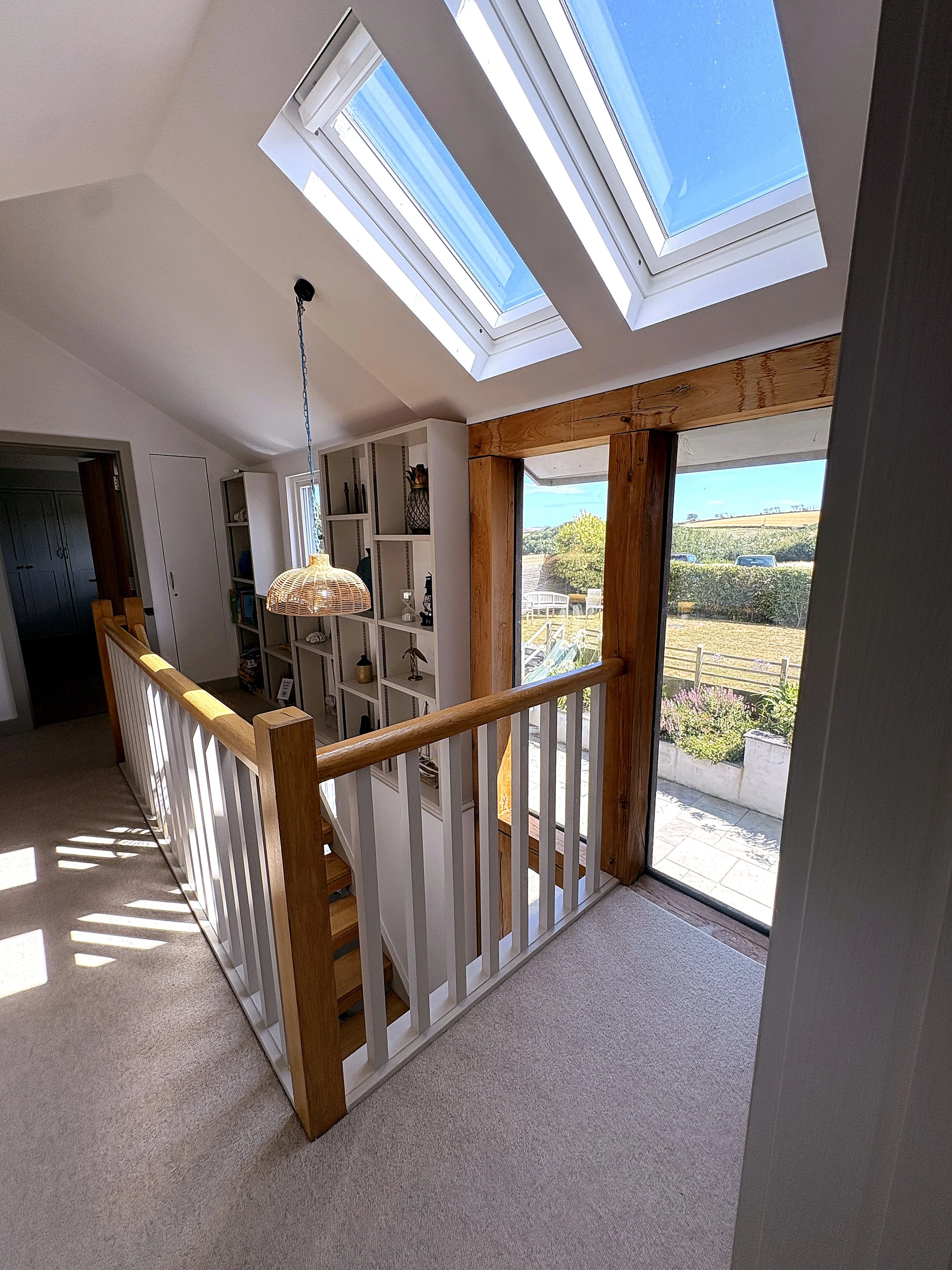 Interior view of a home with a stair railing, large skylight windows on the ceiling, and a sliding glass door that opens to a garden with outdoor furniture and a hedge beyond.