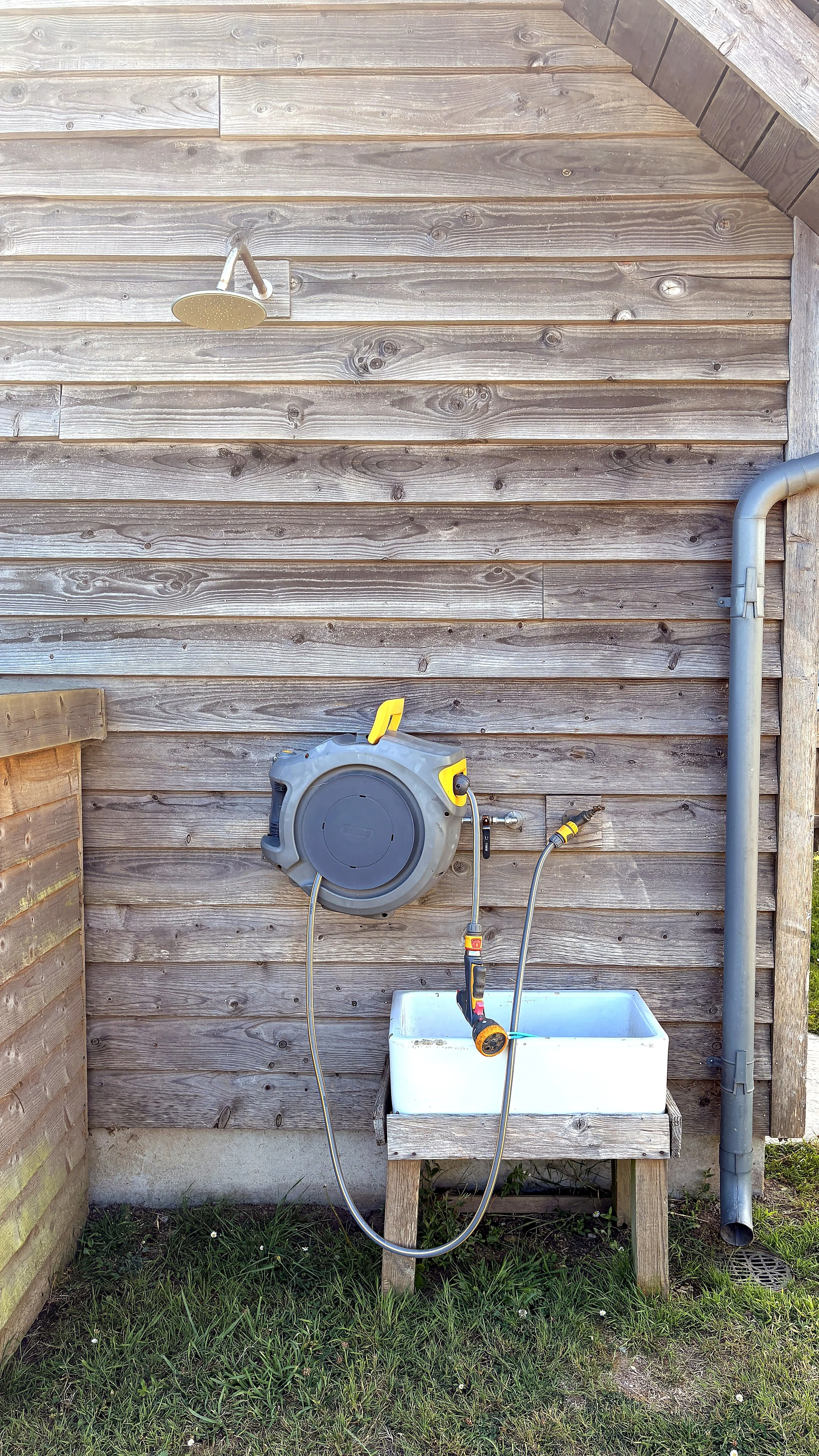 An outdoor wooden wall with a rain shower head mounted on it, a hose reel, a utility sink, and a drainage pipe.