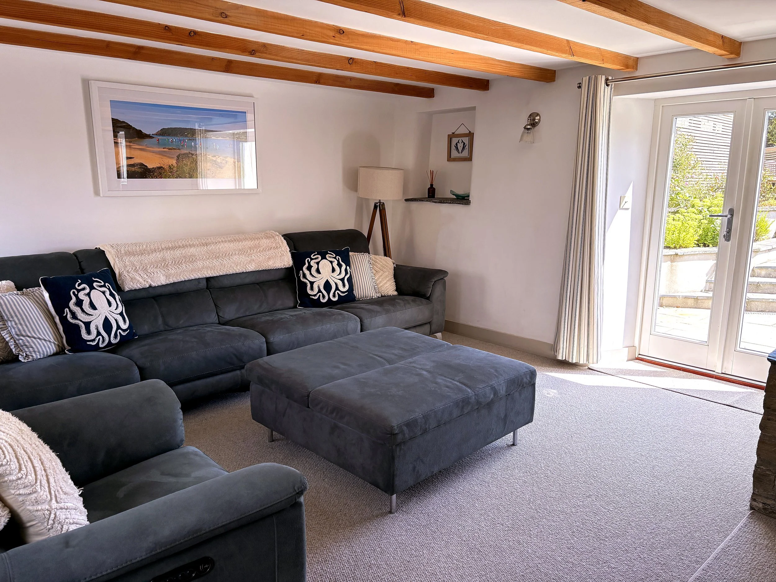 Living room with a dark gray upholstered sofa decorated with navy blue pillows featuring white octopus designs, a large picture window with curtains, and a sliding glass door leading outside. The ceiling has exposed wooden beams, and there is a floor