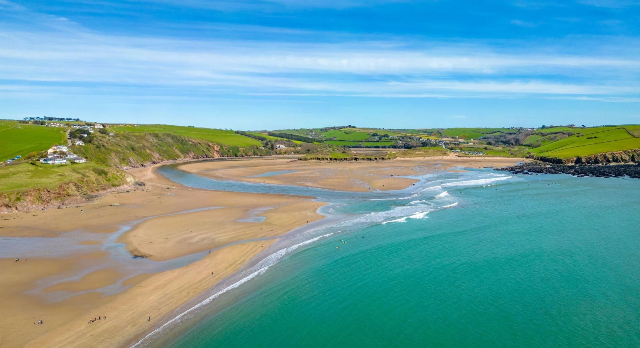 A scenic coastal landscape featuring a sandy beach, turquoise ocean waves, green hills with some houses, and a blue sky with wispy clouds.