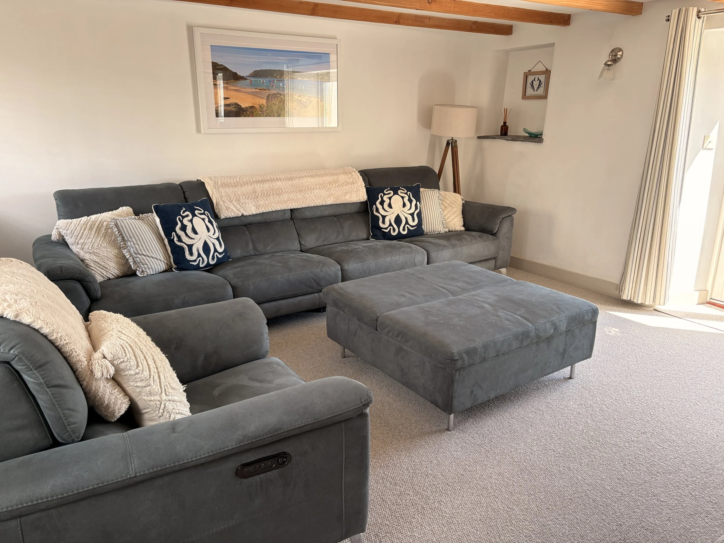 Living room with a gray sofa and pillows, a framed landscape painting of a beach scene, a floor lamp, and a small shelf with decorative items, next to a window with vertical blinds.